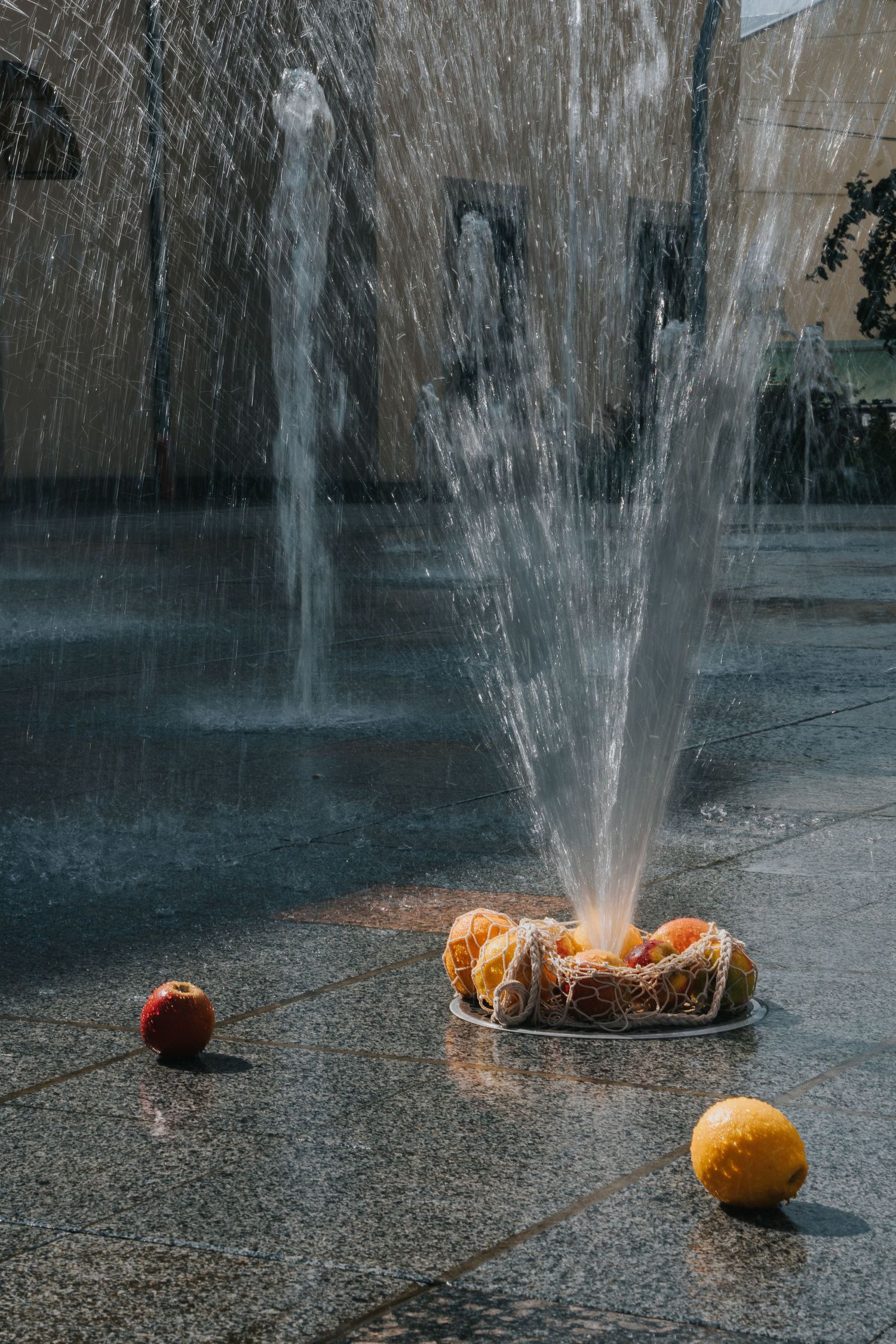 Water fountain spraying over fresh fruits including apples and oranges on a granite plaza.
