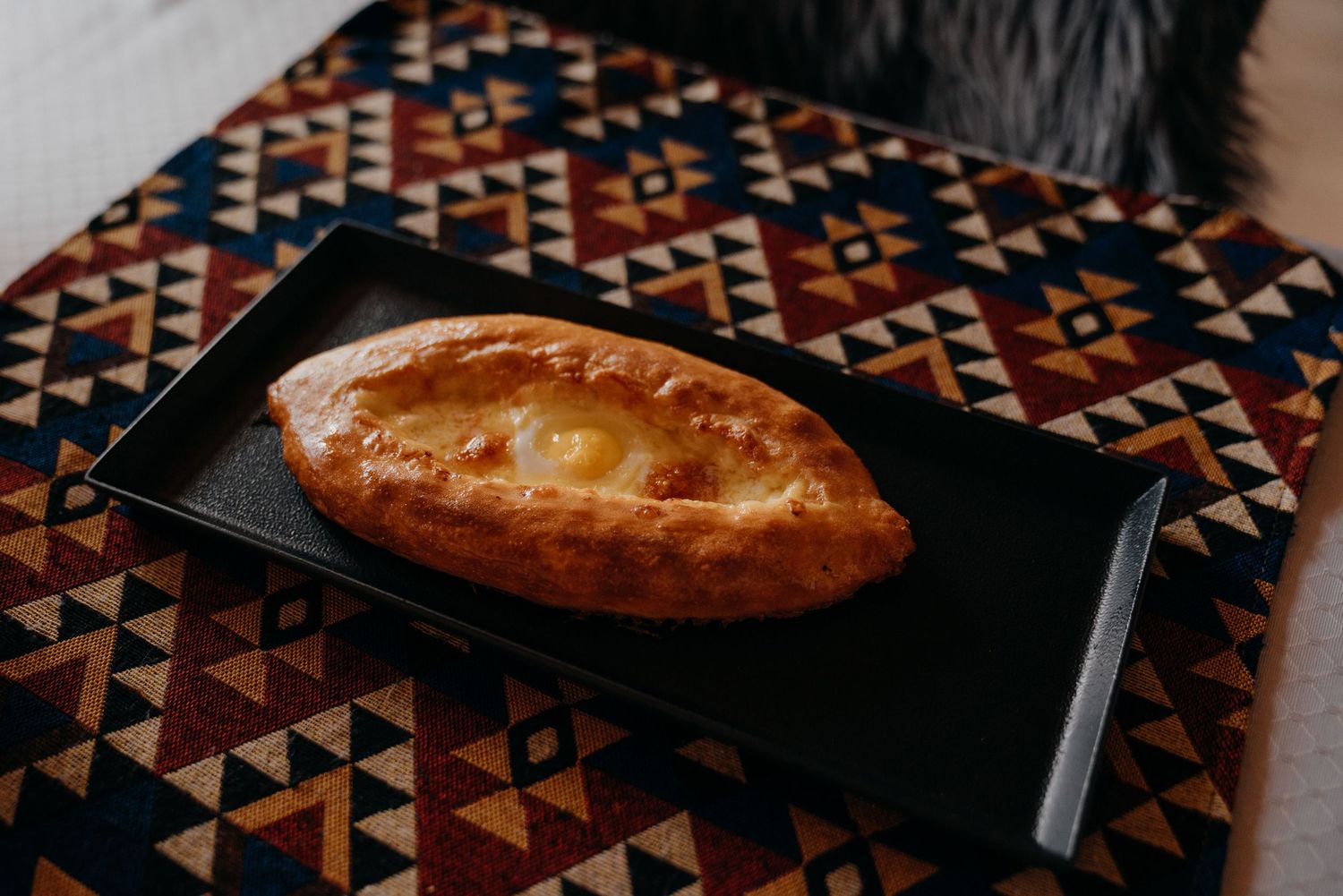 Golden baked khachapuri cheese bread served on a black rectangular plate on a colorful geometric rug.