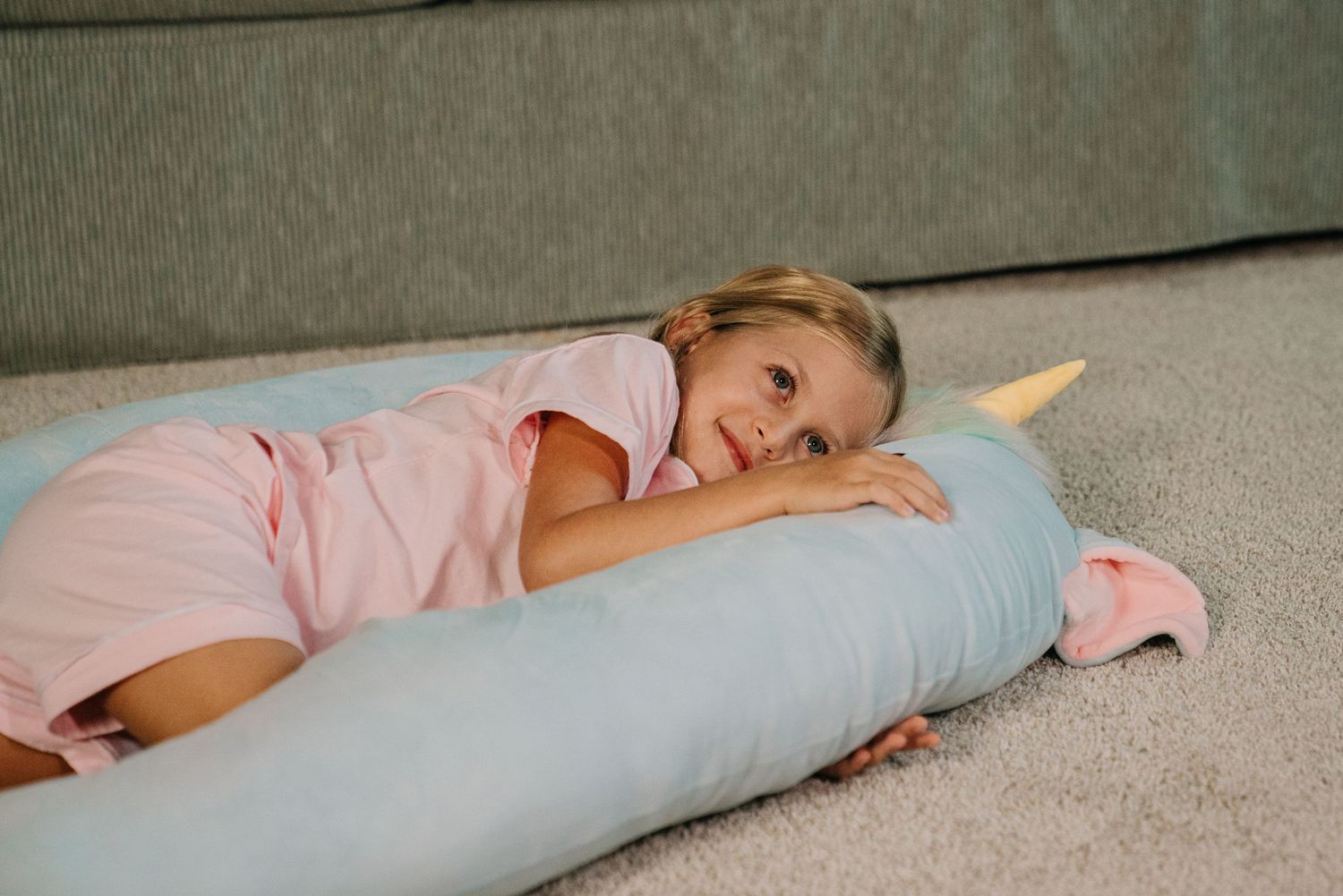 Young girl in pink shirt lying on a light blue unicorn body pillow on a carpeted floor near a sofa.