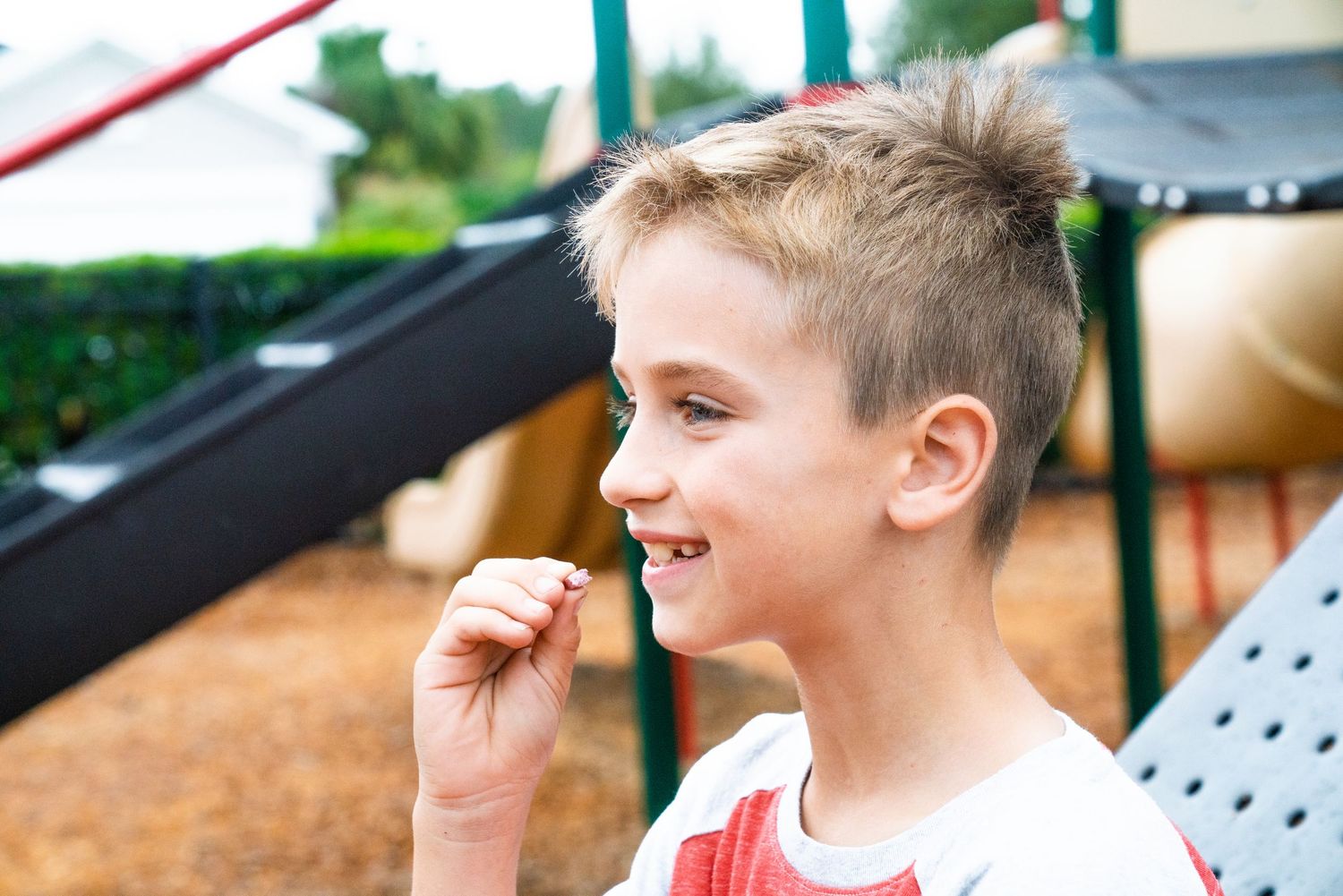 Smiling blonde boy at playground holding small object near mouth with colorful play equipment in background.