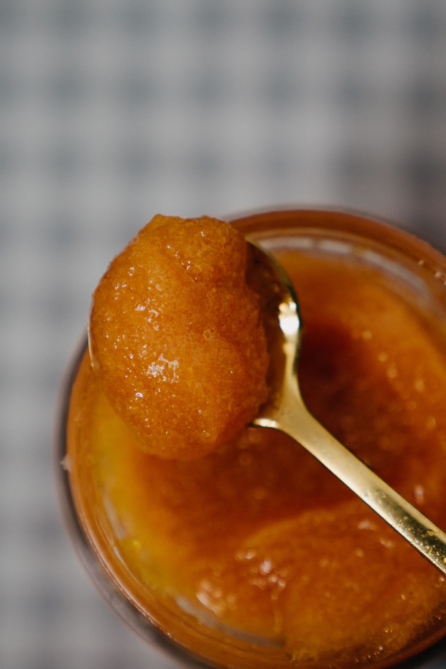 Close-up of a golden spoon scooping thick orange marmalade jam from a glass jar on a checkered cloth.