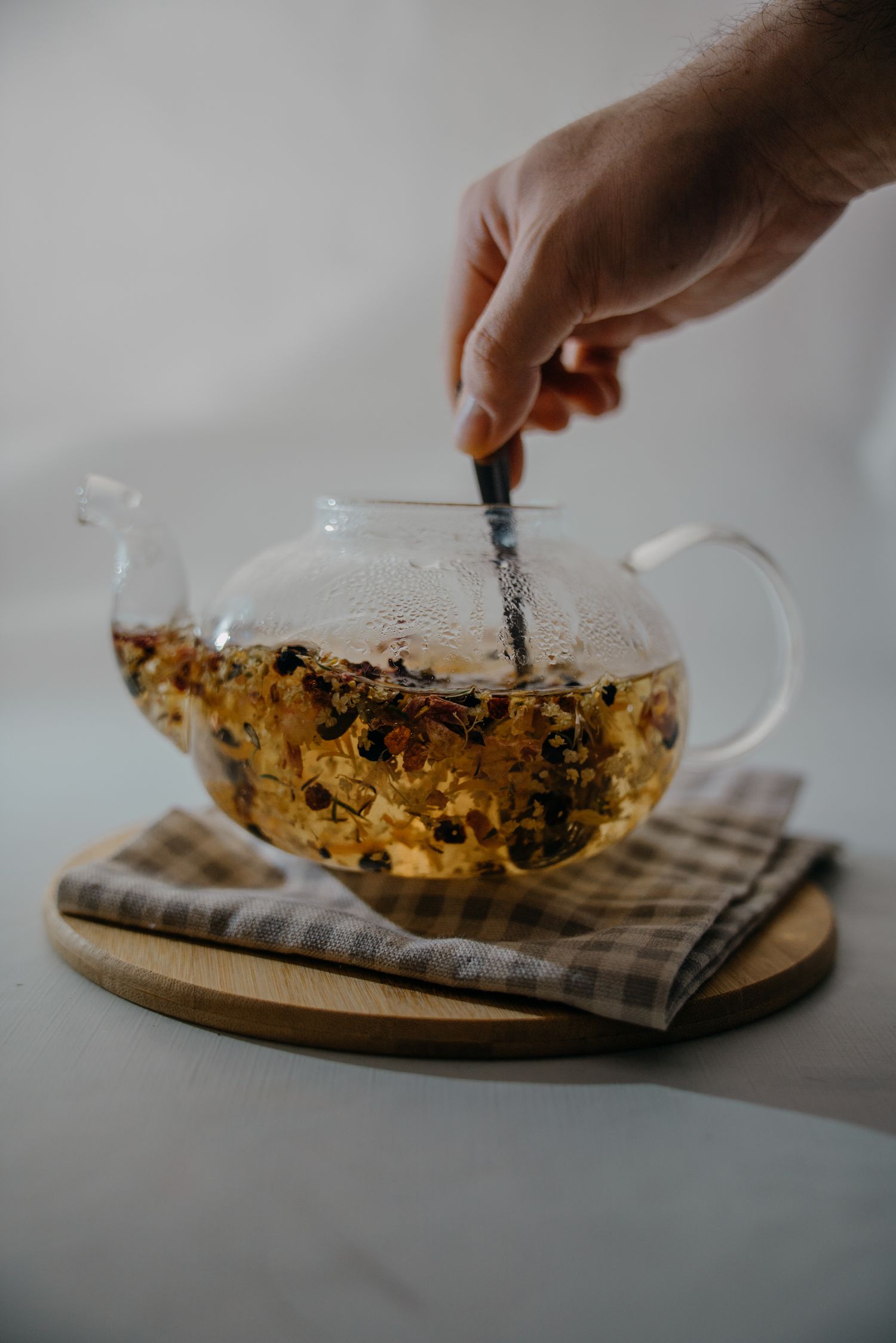 Hand stirring herbal tea in a clear glass teapot on a wooden board with a checkered cloth.