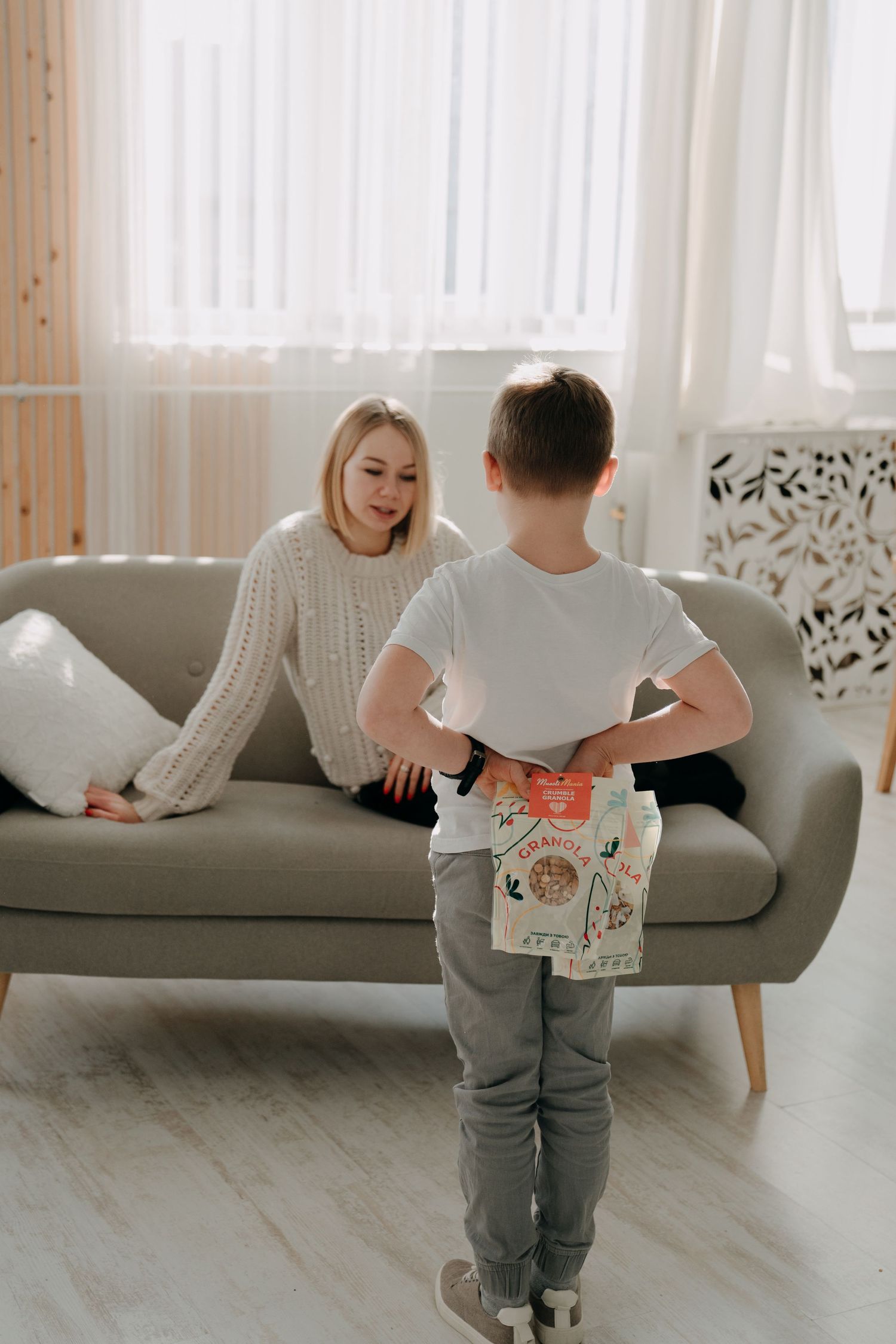 Young boy hiding a granola gift bag behind his back as a surprise for a woman sitting on a gray sofa.