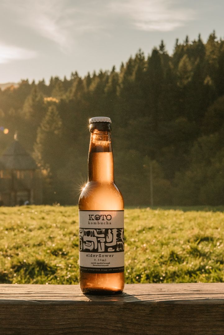 KOTO Kombucha elderflower bottle on wooden surface with golden sunset light, green meadow and pine forest background.