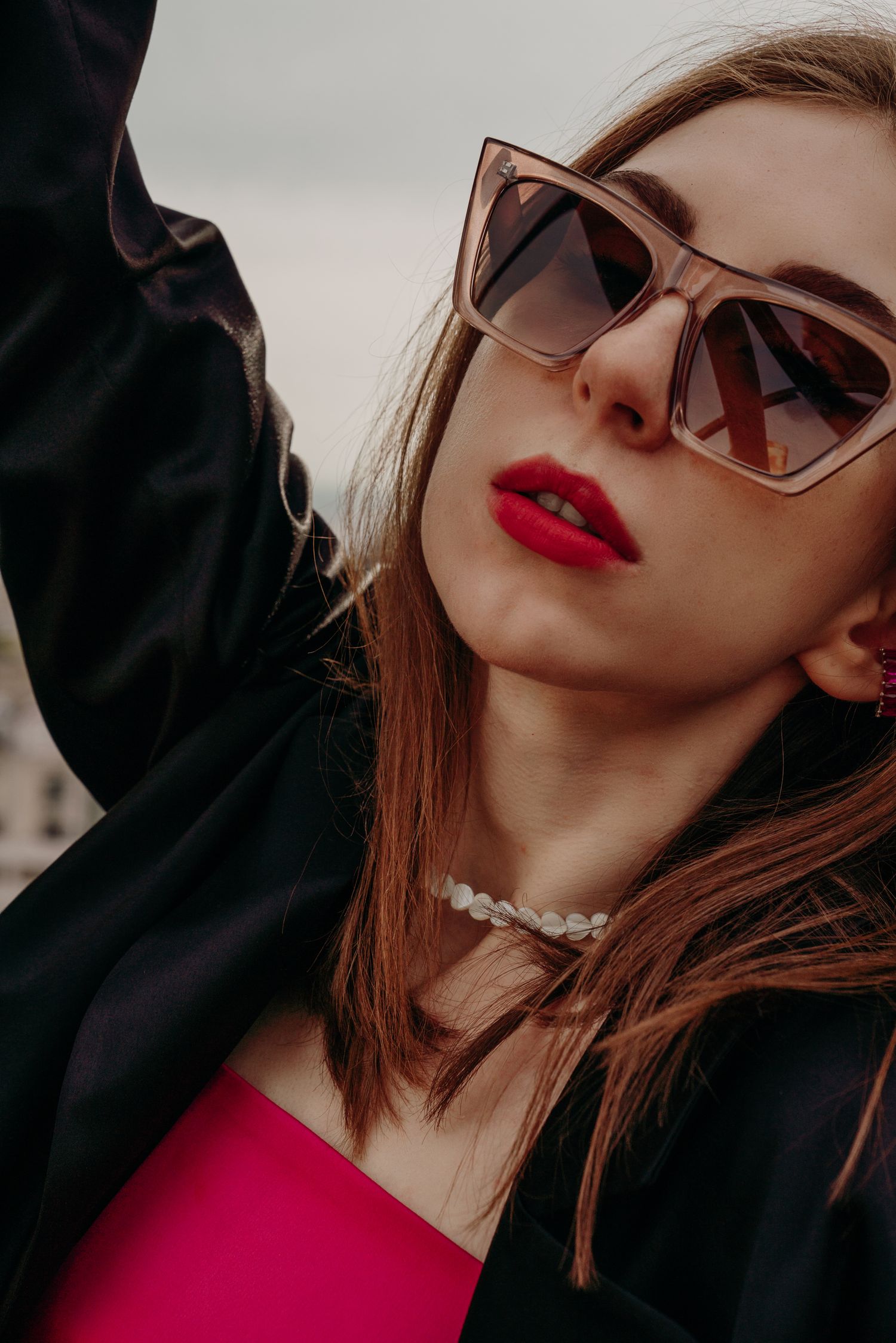 Close-up of stylish woman wearing oversized cat-eye sunglasses and pearl necklace with red lips outdoors.