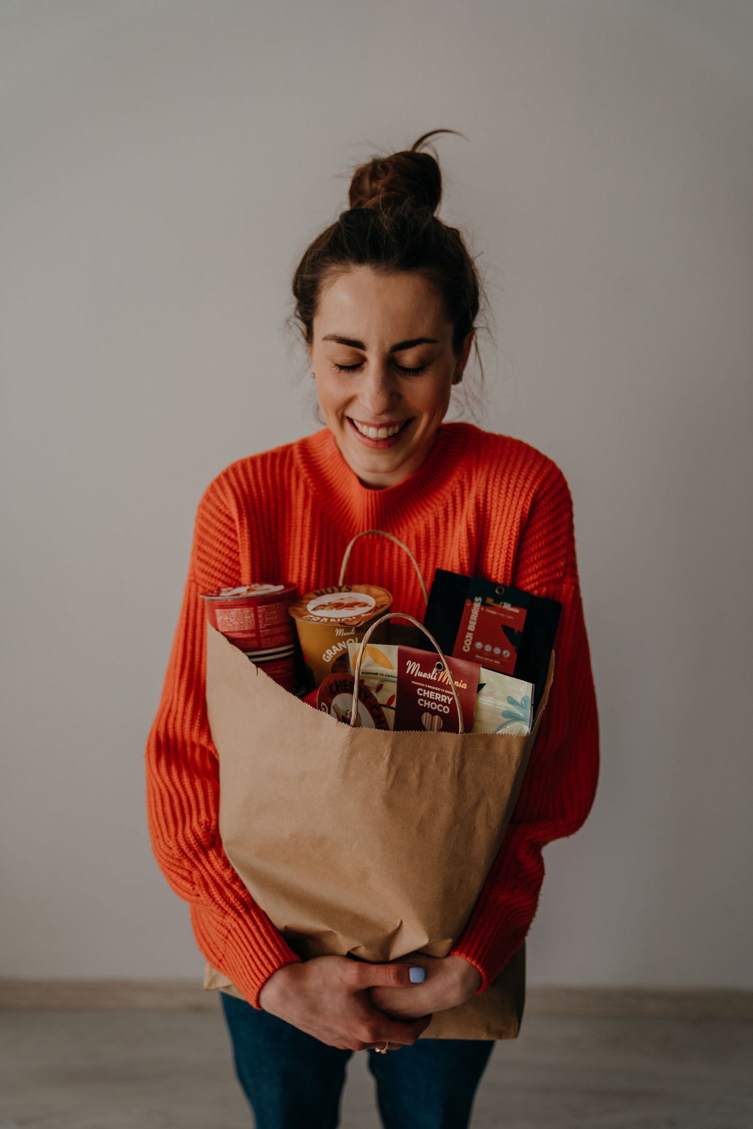 Smiling woman in orange sweater holding a brown paper bag filled with gourmet food products and snacks.