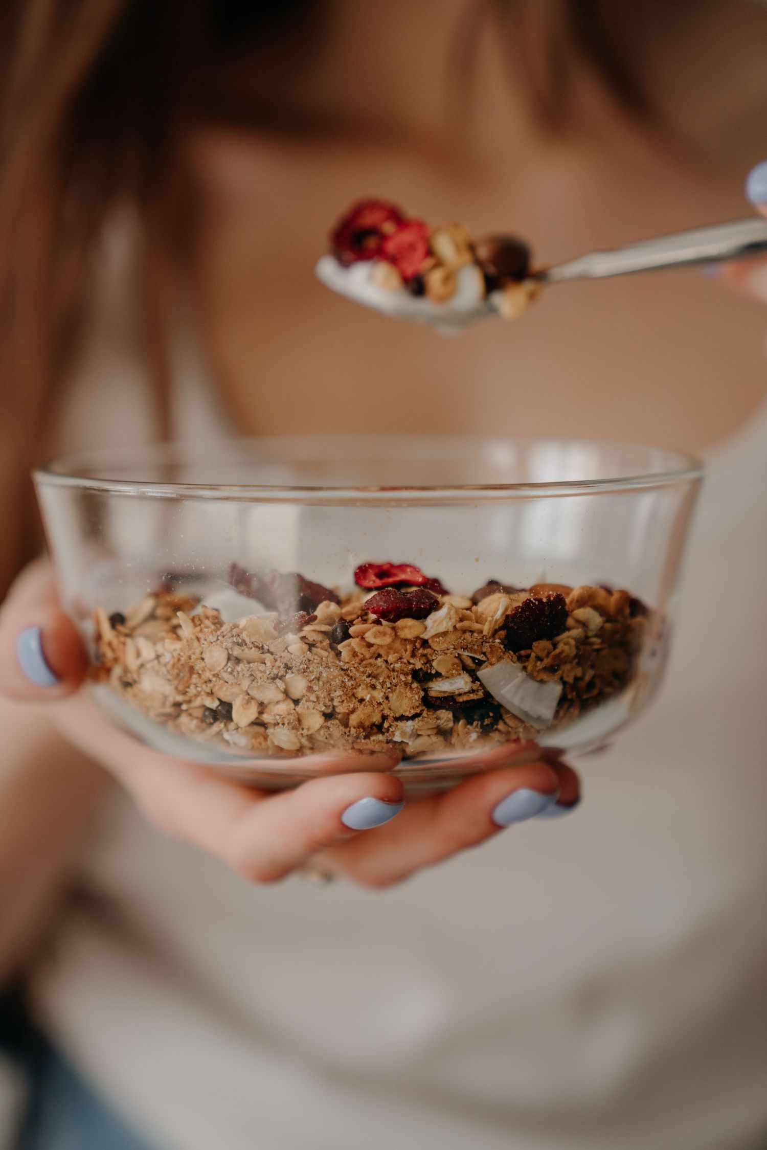 Woman holding glass bowl of granola with dried berries, lifting spoonful of crunchy muesli mix.