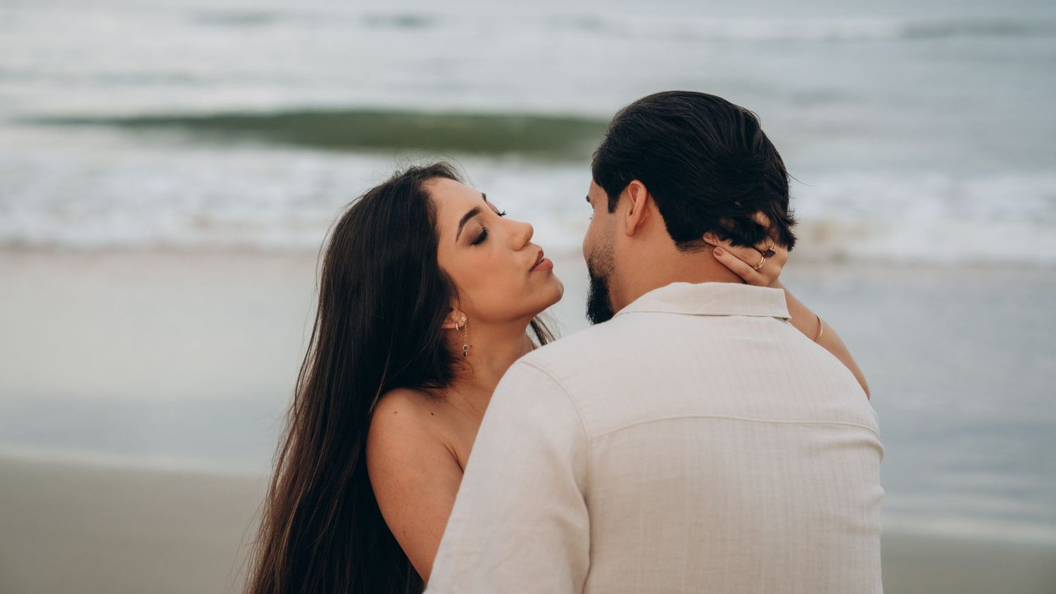 Panoramic beach scene of a couple embracing with ocean waves in the background.