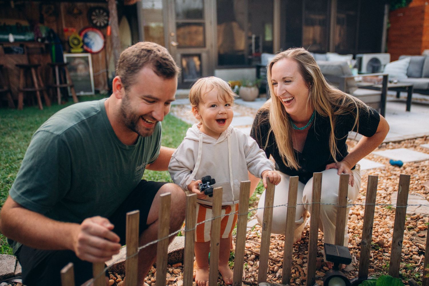 Family members interact playfully near a wooden fence in a backyard setting.