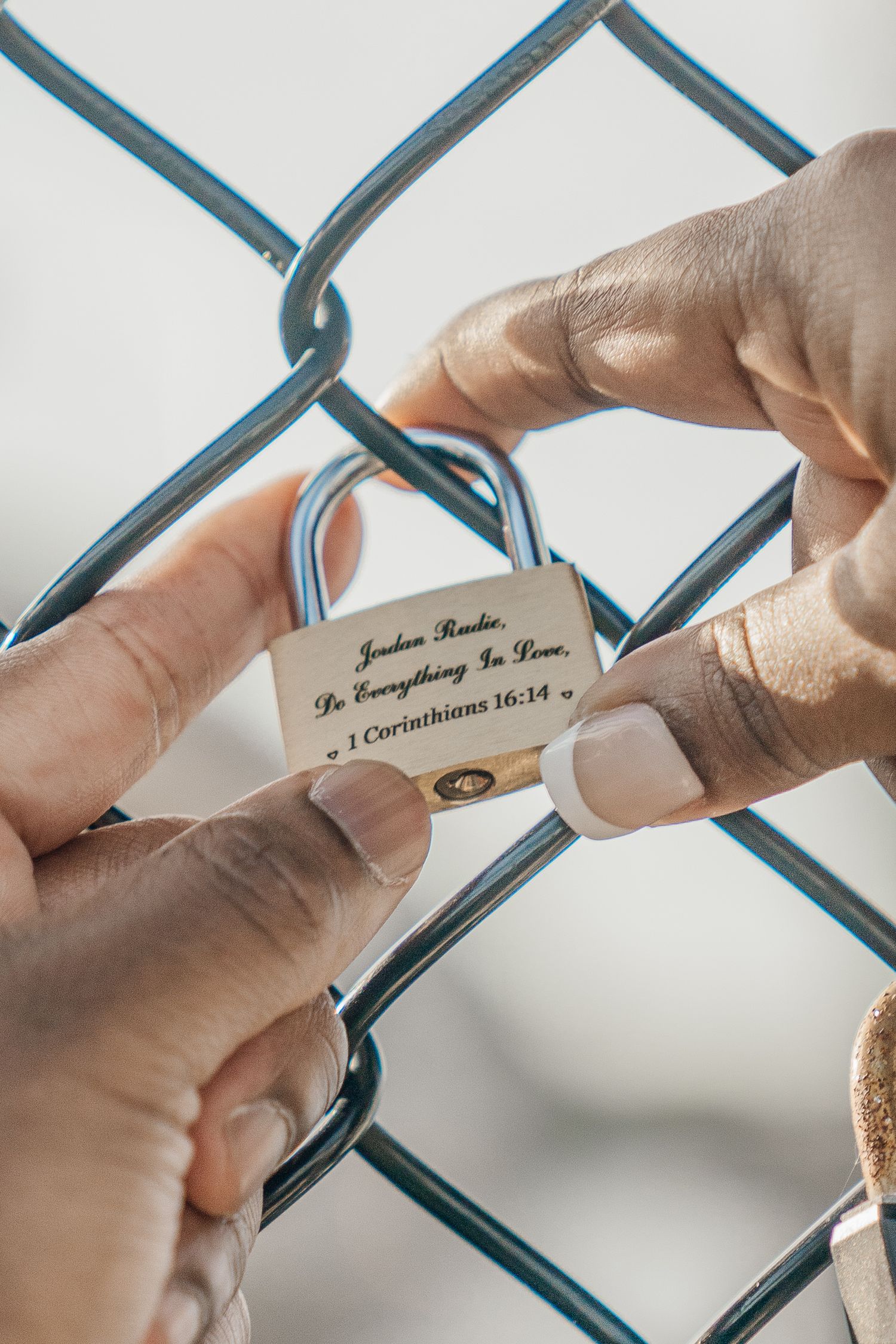 Hands holding a small white lock with a biblical inscription against a chain-link fence.