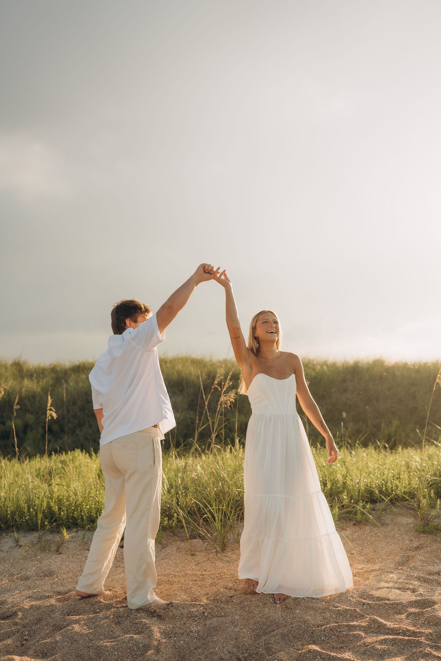 A couple in white clothing dances together on a hilltop during golden hour.