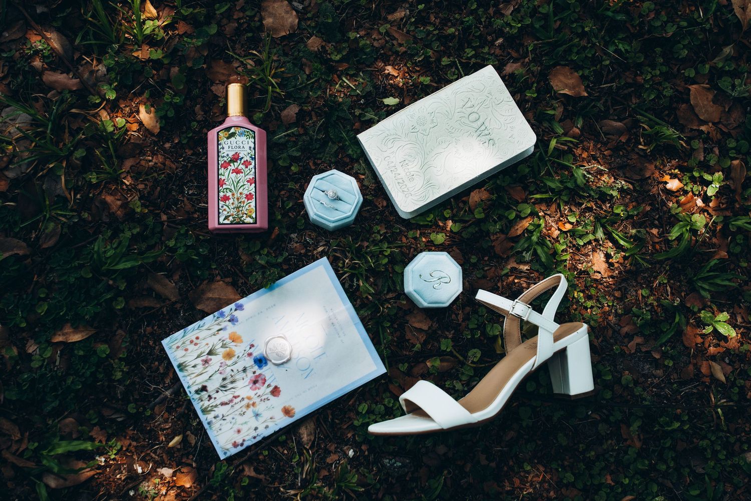 Flat lay of bridal accessories including white heels, floral clutch, perfume bottle, and blue hair pieces on grass.