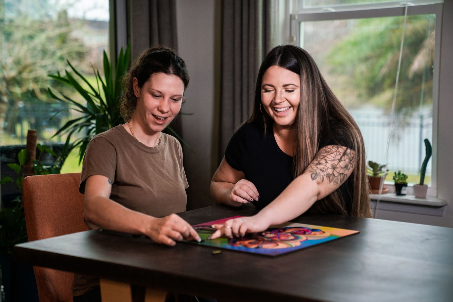 Two women smiling and playing a colorful board game together at a wooden table near a bright window.