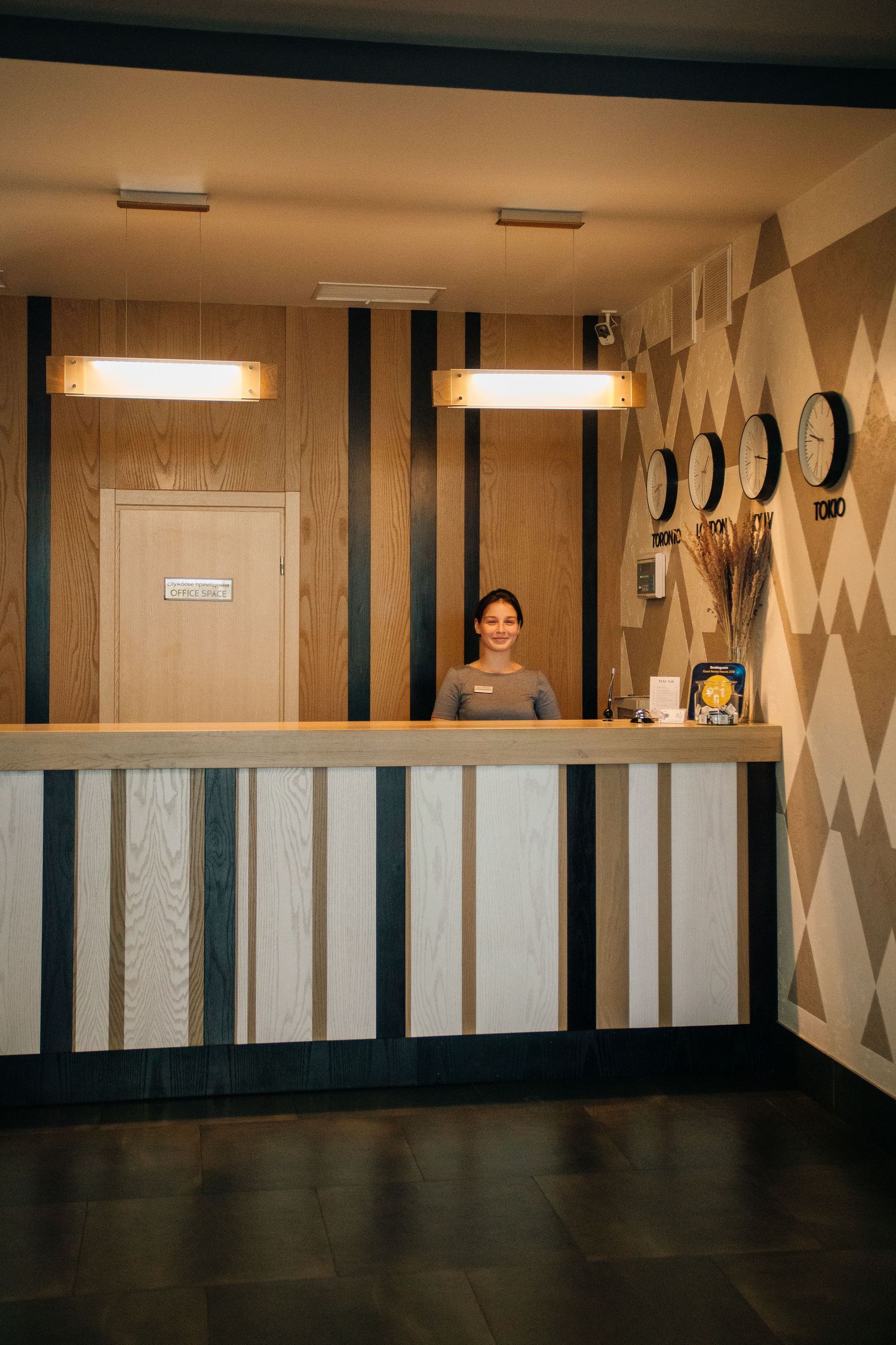 A smiling hotel receptionist stands behind a modern wooden front desk with clocks on the wall.