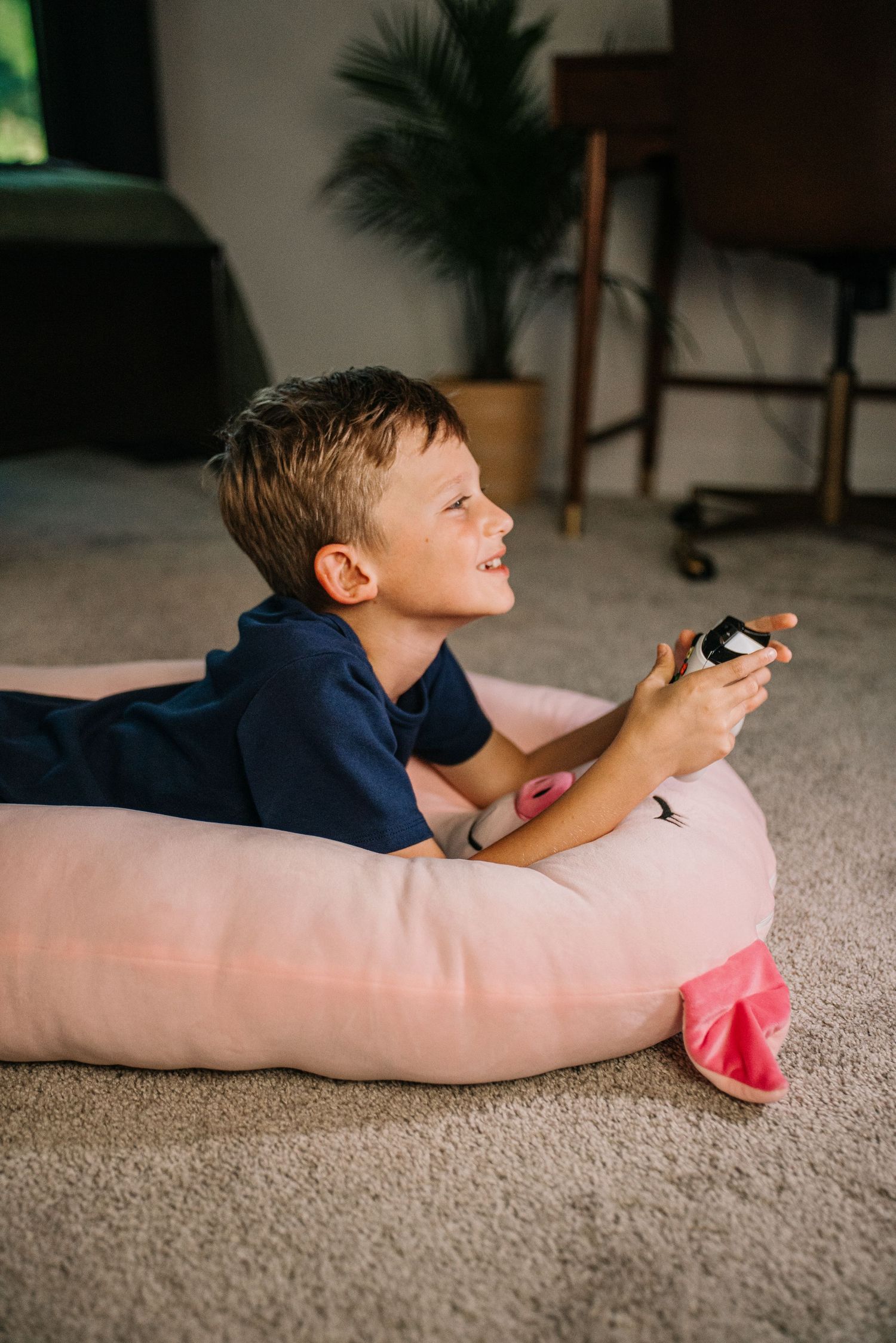 Happy boy playing video games with a controller while lying on a large pink stuffed animal pillow.