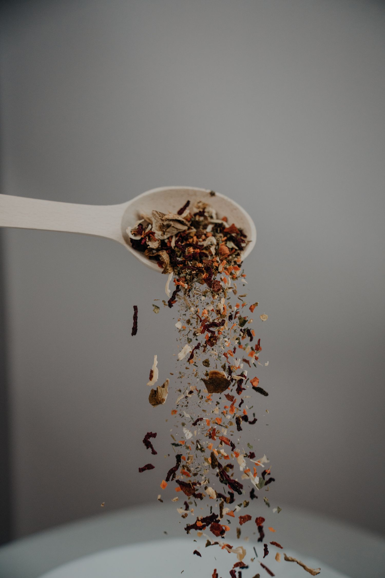 Dried herbal tea blend falling from a white spoon against a gray background.