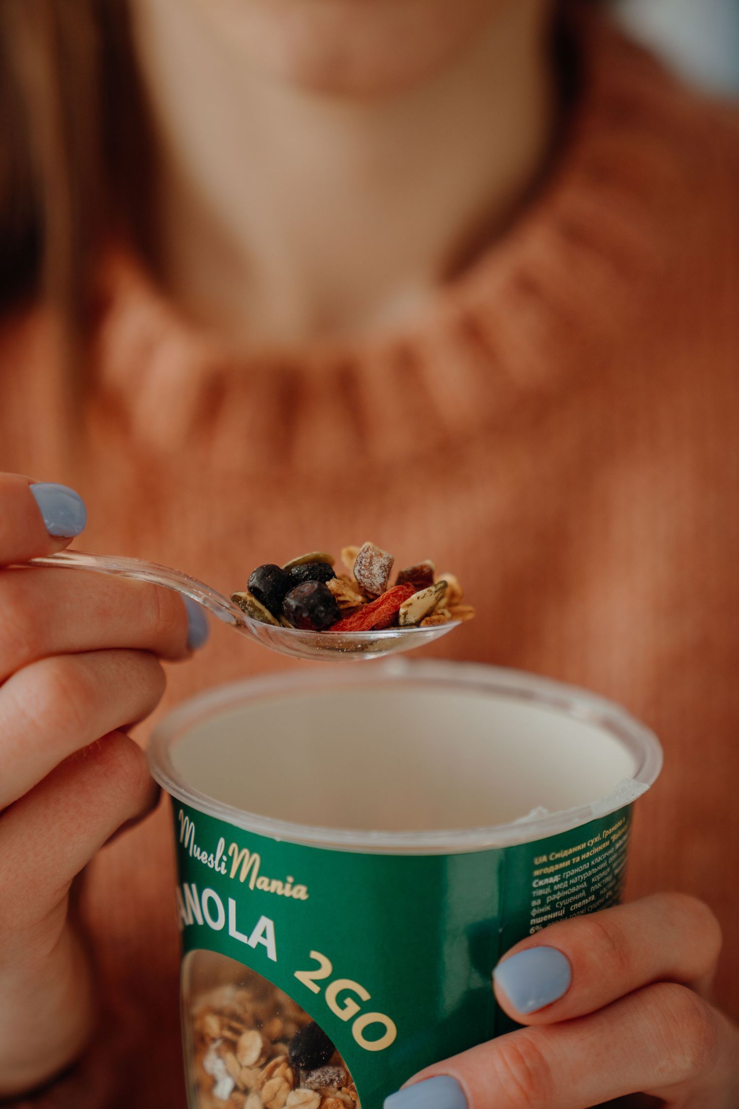 Woman holding a spoon of granola over a MuesliMania Granola 2GO cup, wearing an orange sweater with blue nail polish.