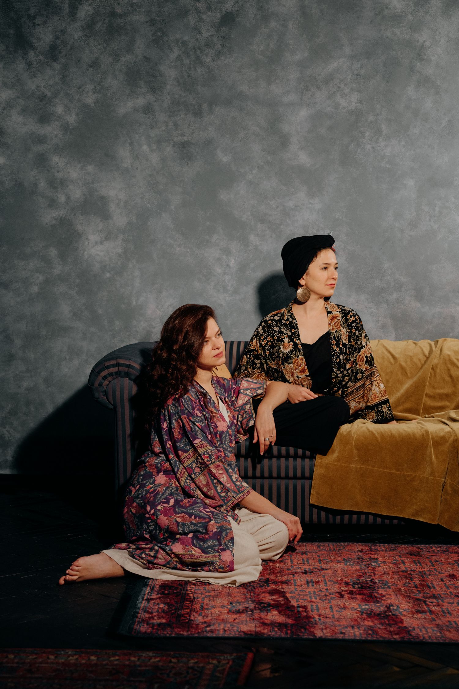 Two elegant women in floral robes pose in a moody studio, one seated on floor, other on gold velvet sofa.