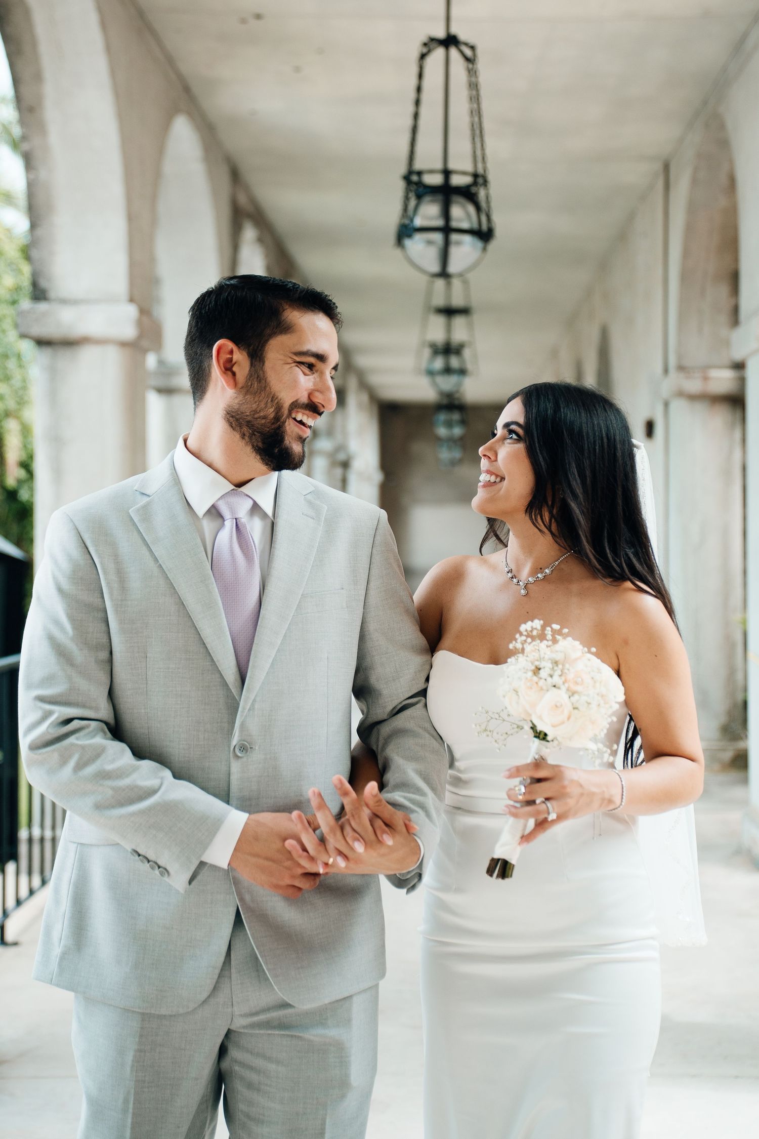 A newlywed couple shares joyful candid moments together in an elegant outdoor colonnade with hanging lanterns.