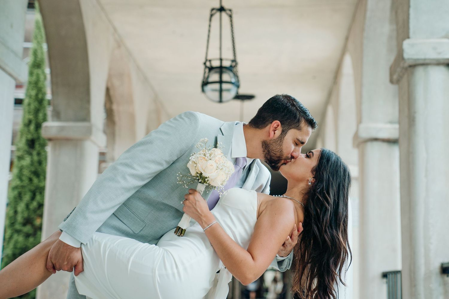Bride and groom share a romantic dip kiss outdoors, with the groom in a light suit holding a white bouquet.