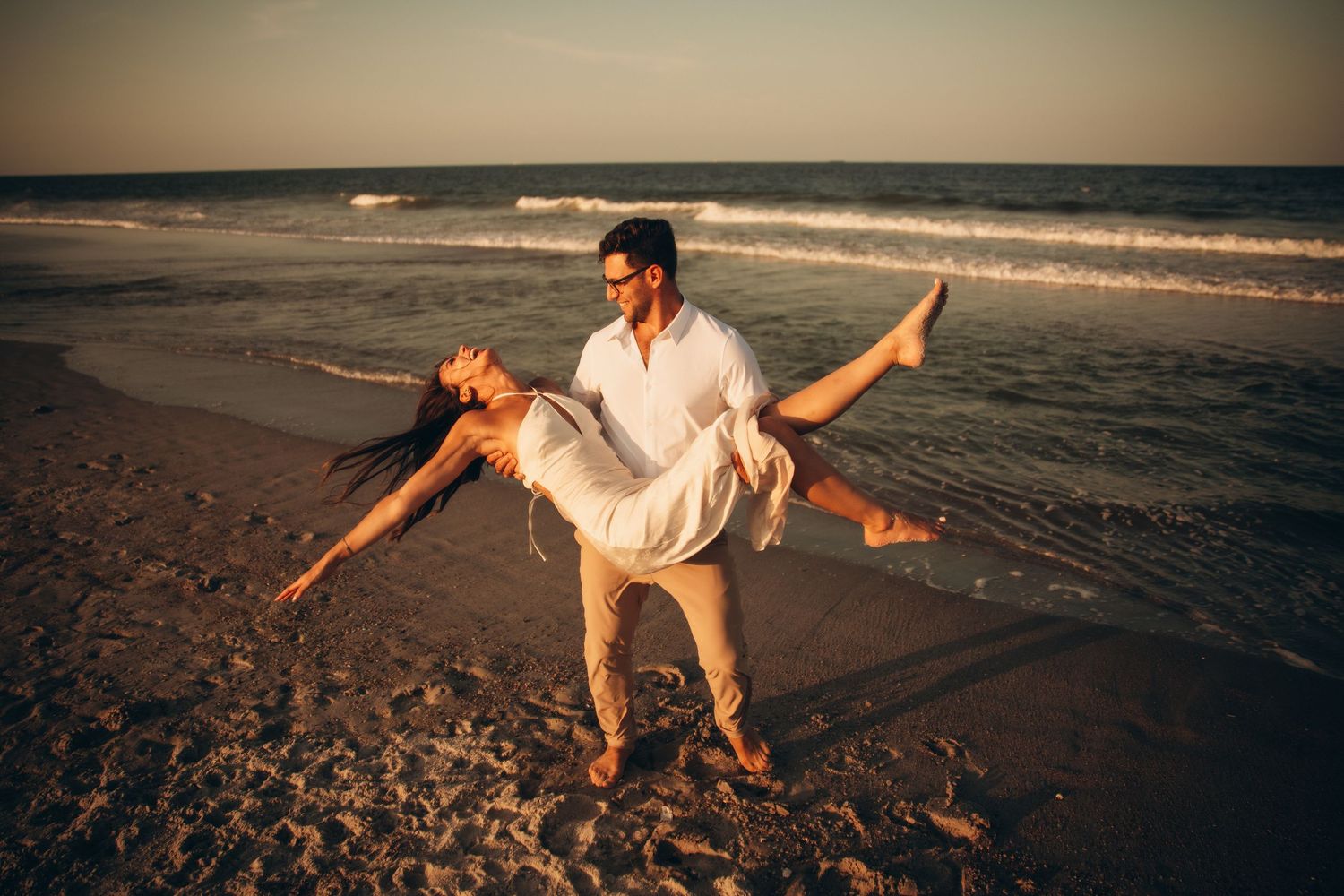 Couple dancing romantically on a sandy beach at sunset, man lifting woman in white dress near ocean waves.