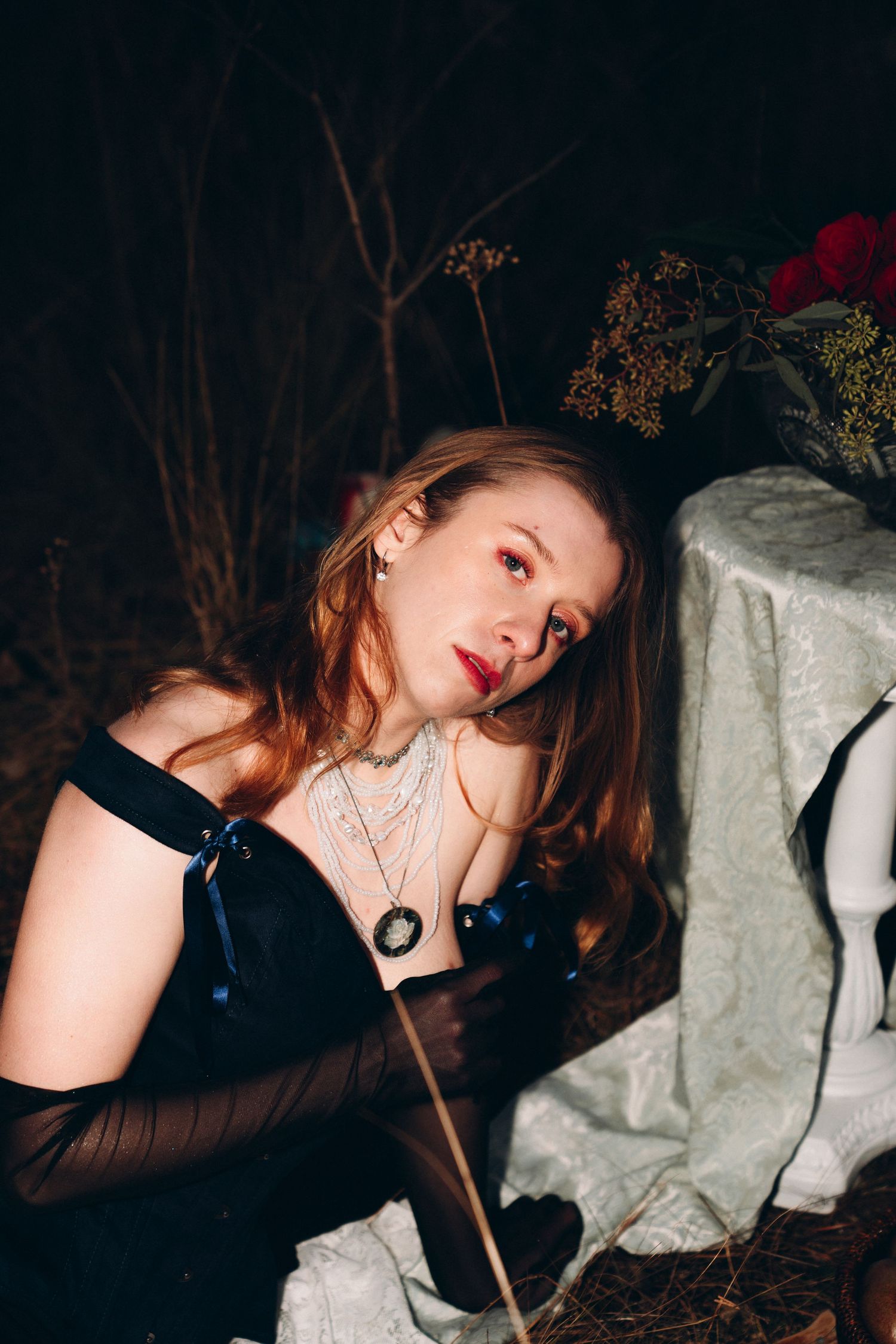 Woman in black corset gown with sheer gloves and cameo necklace poses dramatically beside lace-covered table at night.