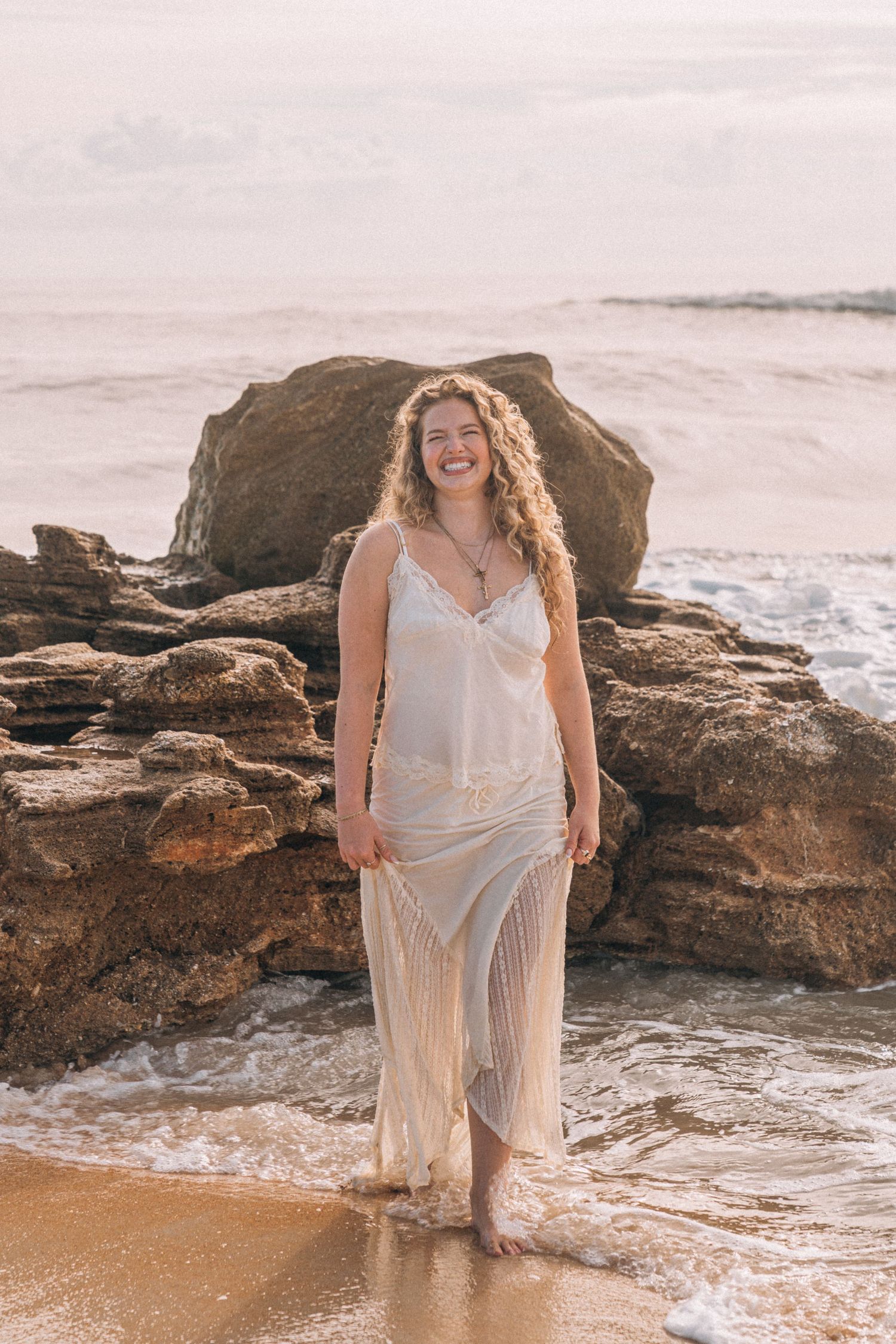 Smiling woman in white fringe dress standing barefoot on sandy beach with rocky shoreline and ocean waves behind her.