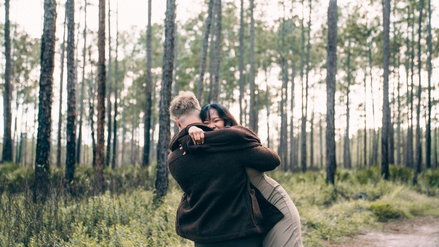 A romantic couple embraces in a sunlit forest with tall pine trees creating a dreamy atmosphere.