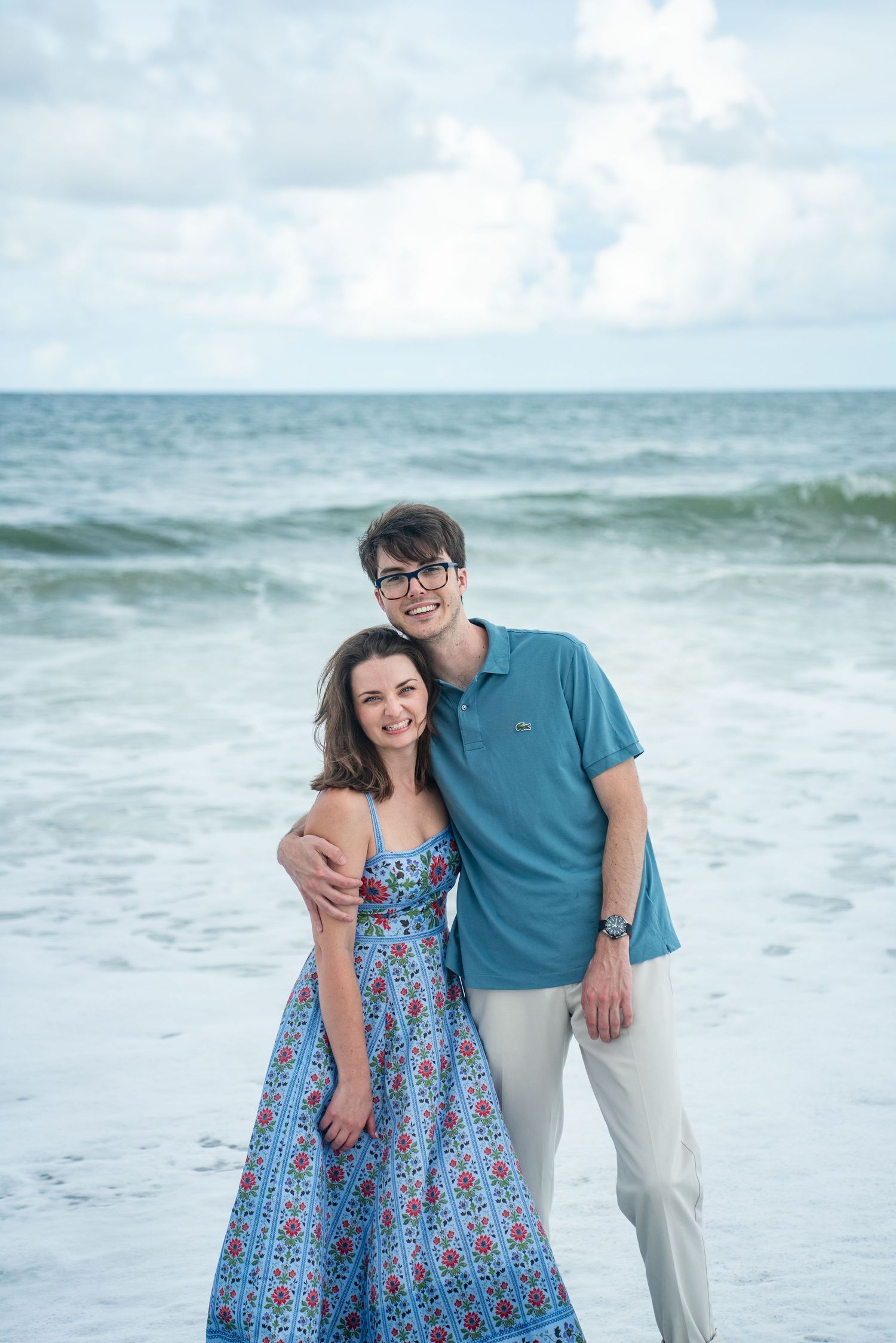 A couple stands together smiling on the beach with ocean waves in the background.