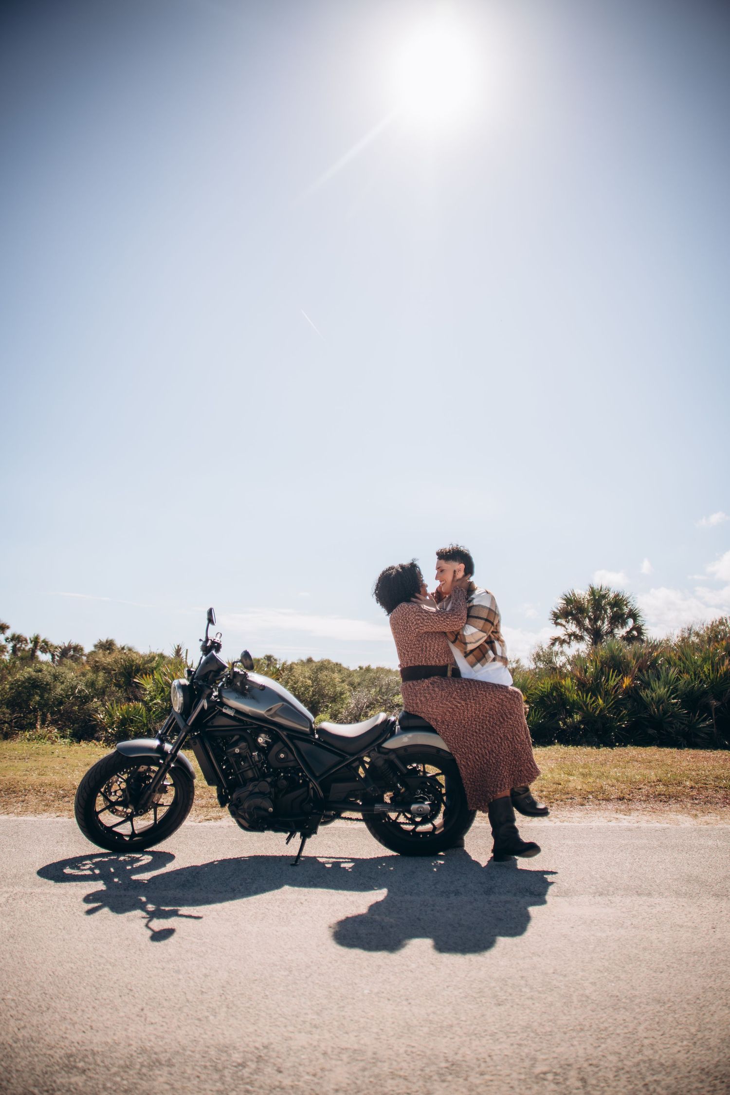 A couple sits together on a parked motorcycle on a sunny day with palm trees visible in the distance.