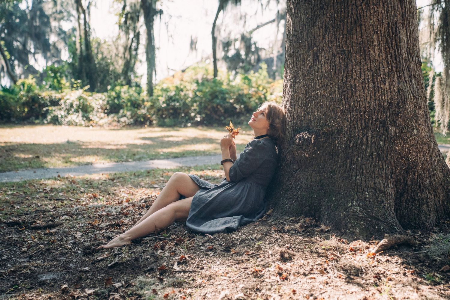 Someone in a black dress relaxes against a large tree trunk in a sunlit forest setting.