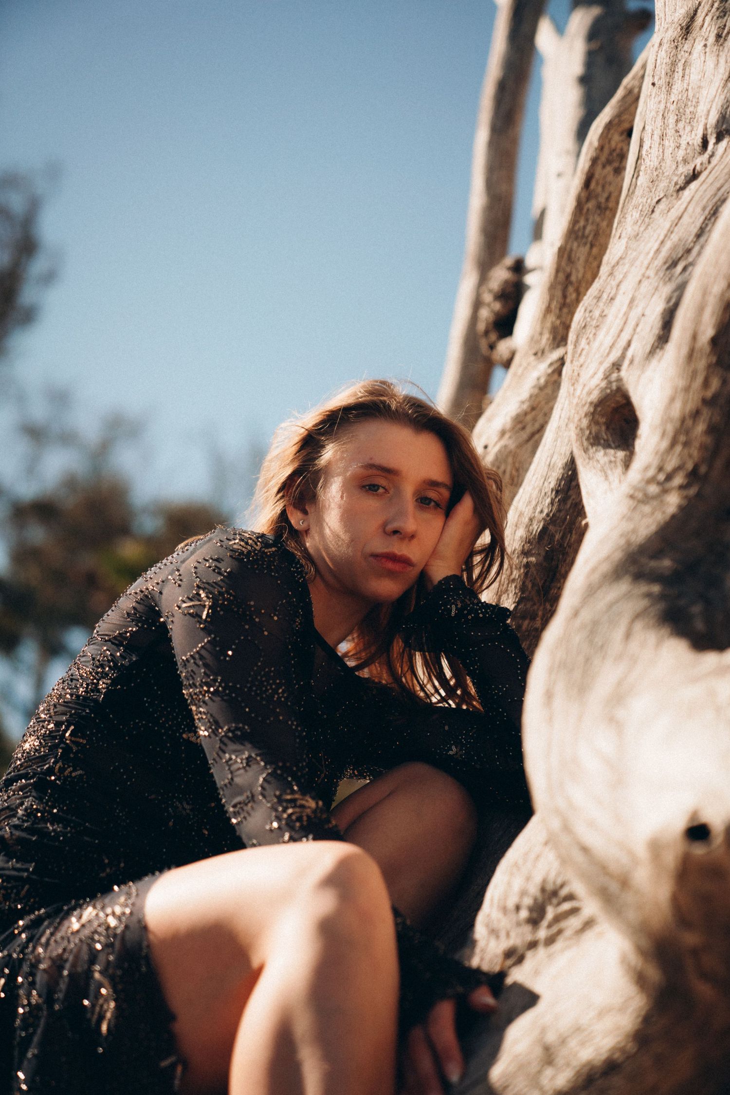 A figure in a black dress rests against weathered driftwood on a beach in dramatic lighting.