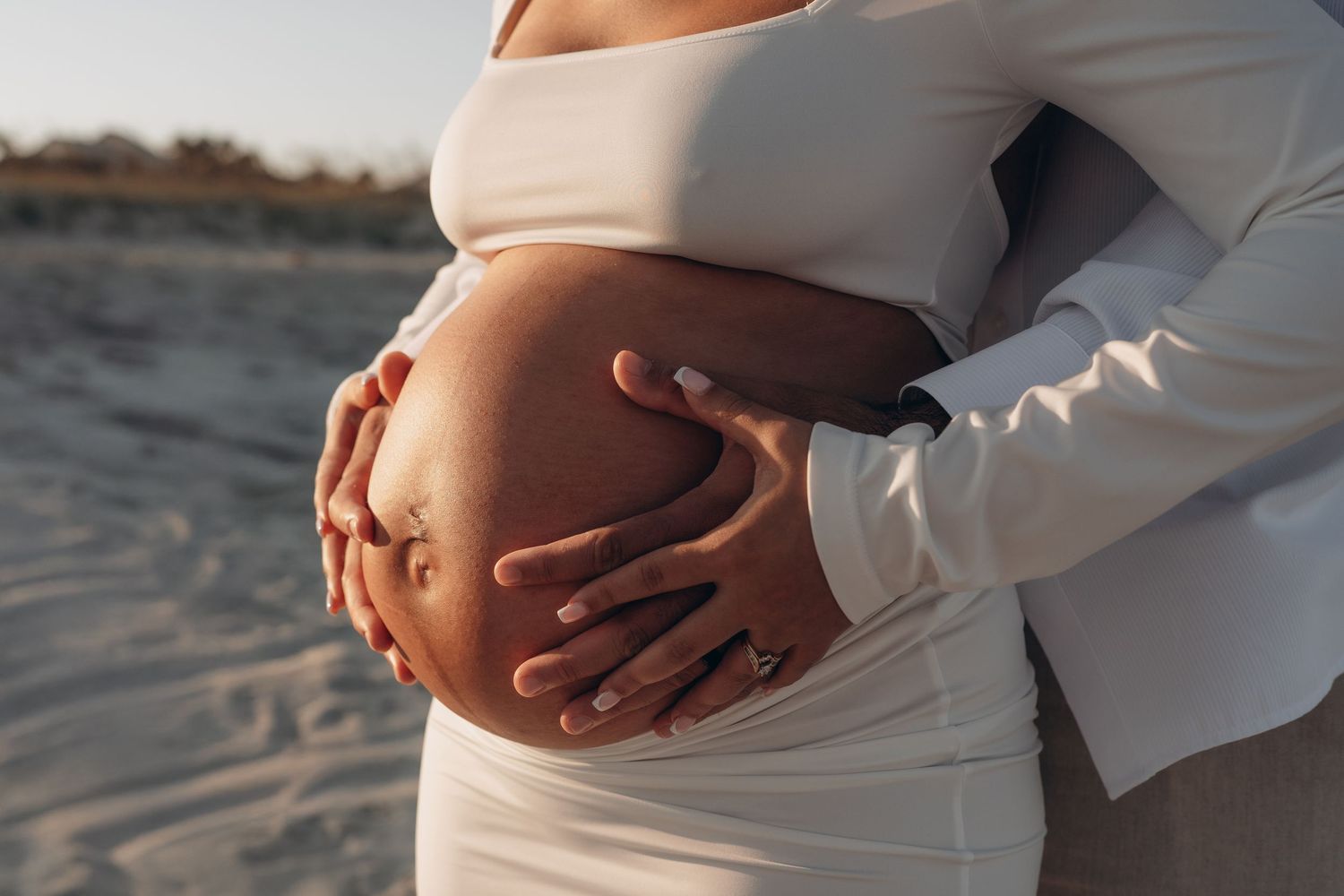 Close-up view of a pregnant belly being cradled on a sunlit beach.