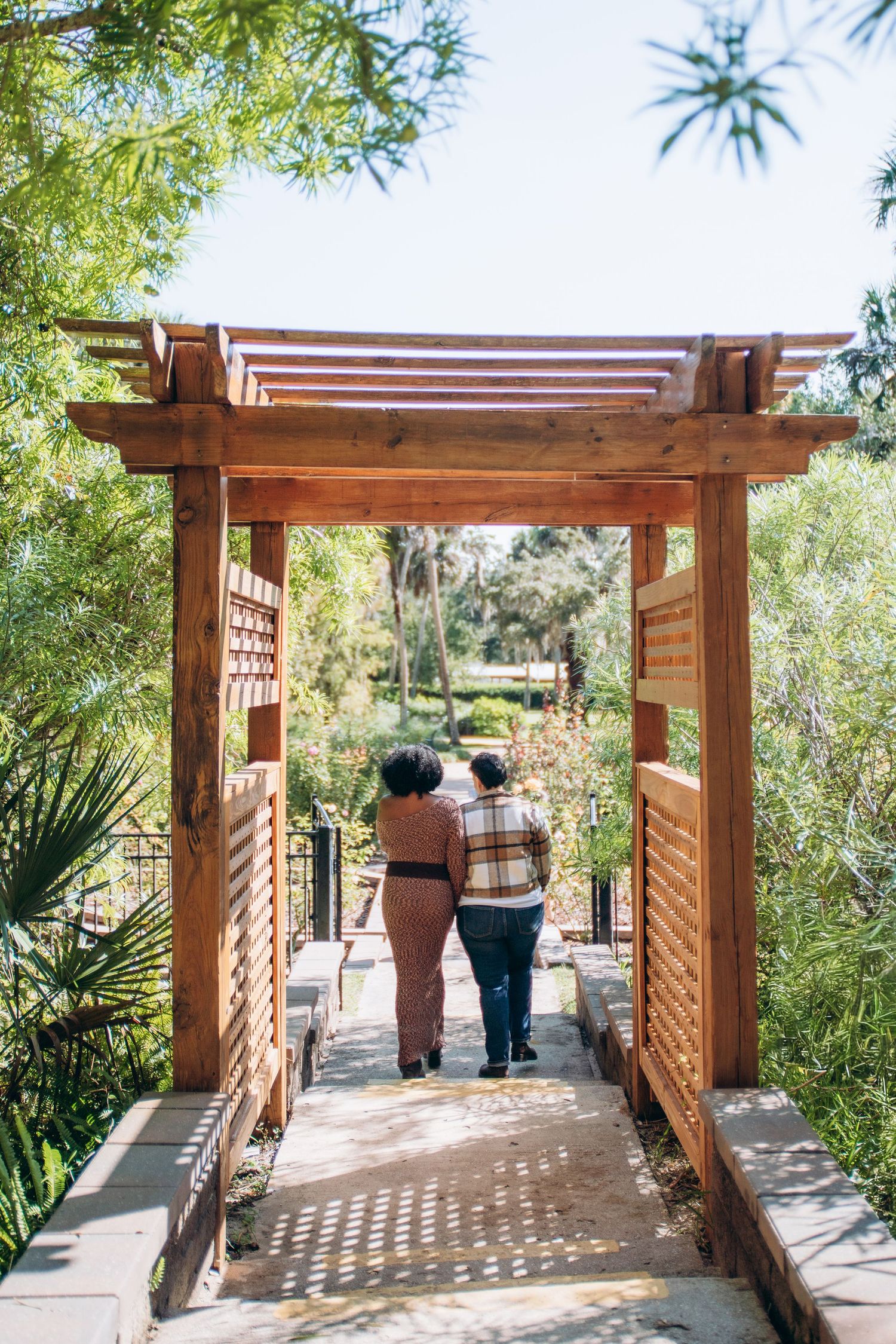 A wooden pergola walkway with lattice sides creates dappled shadows in a lush garden setting.