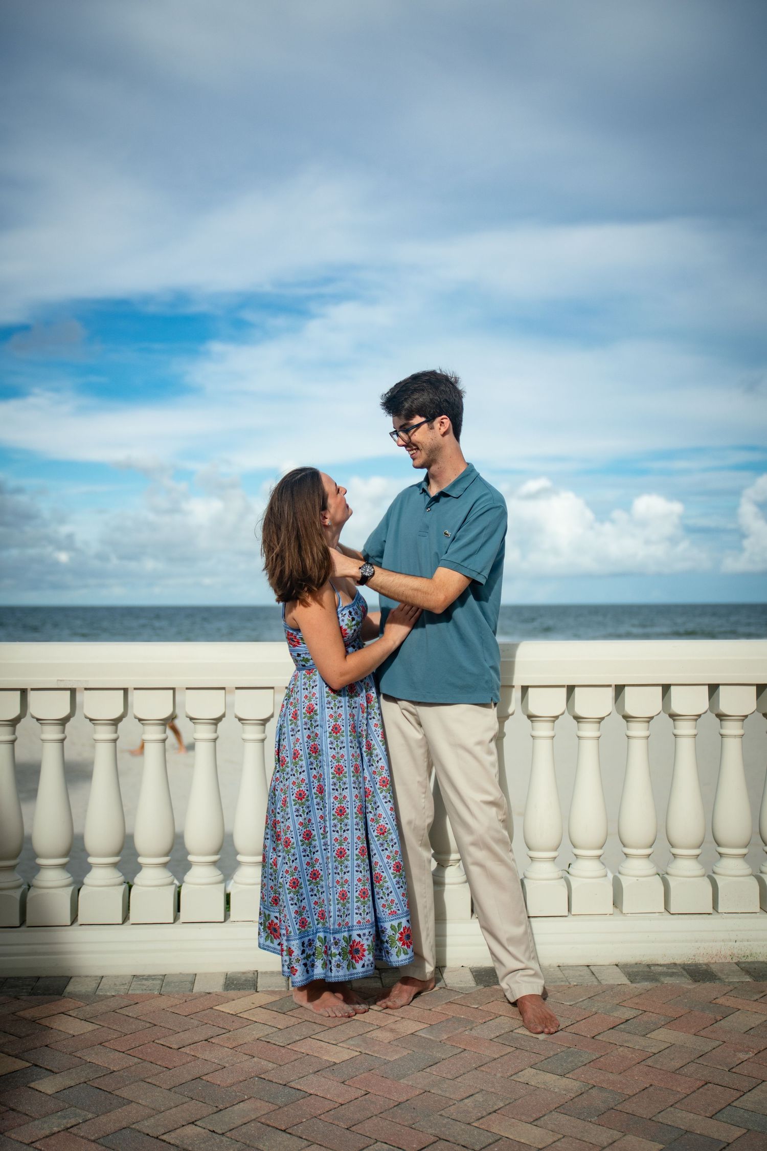 A couple embracing on a beachside balcony with ornate white railings against a bright blue sky.