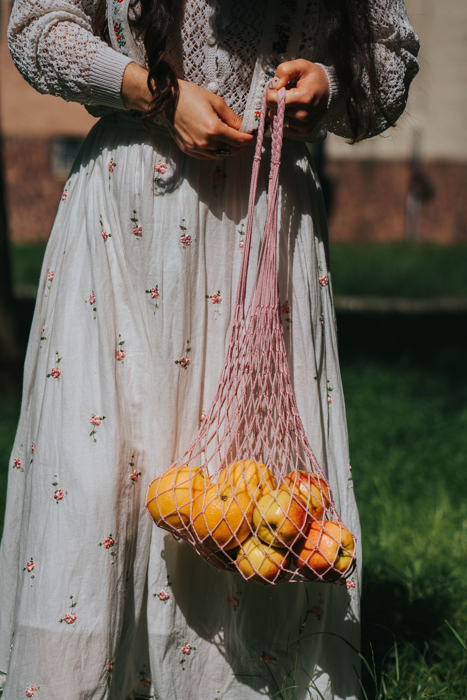 Woman in floral dress holding pink mesh bag filled with oranges and lemons outdoors.