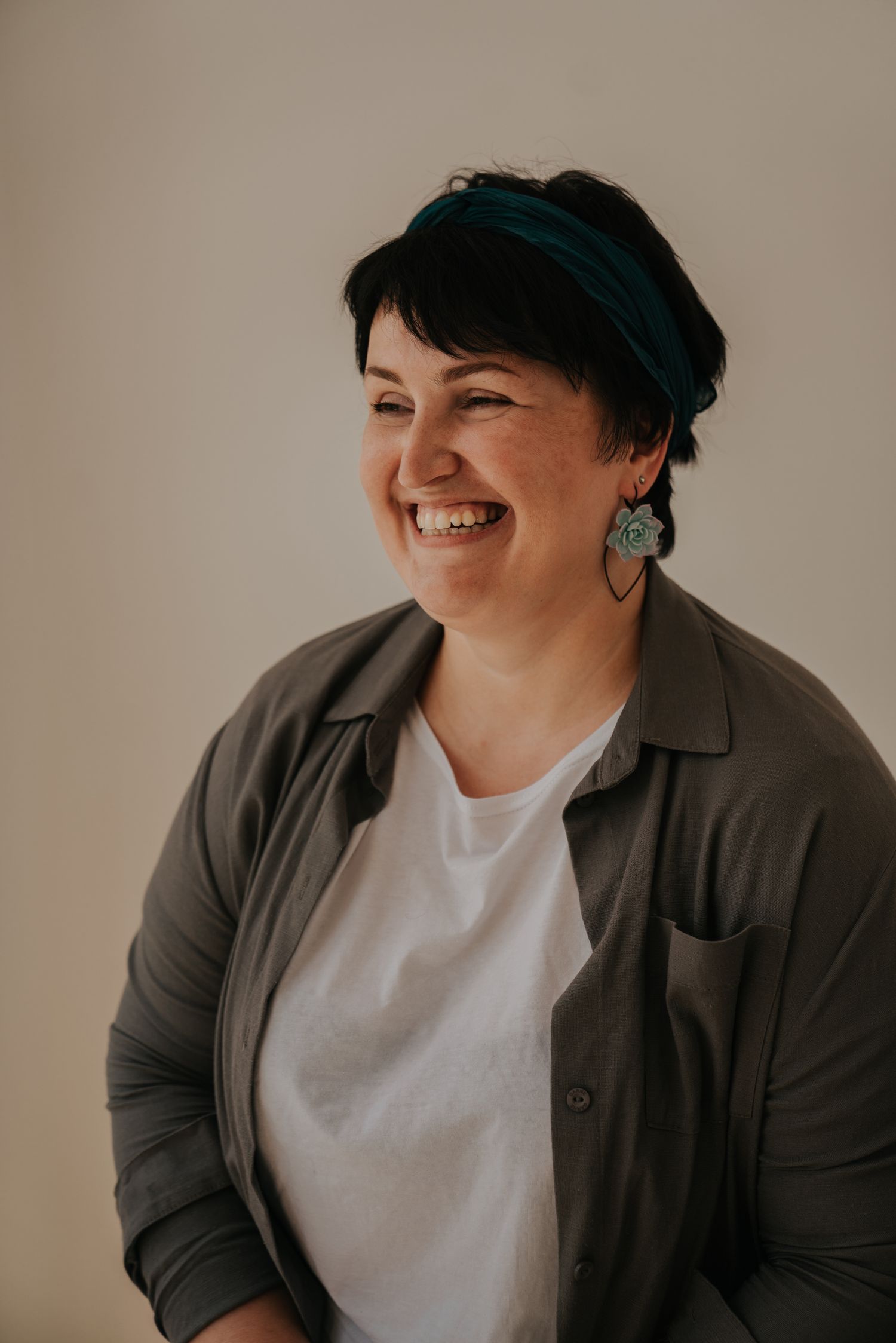 Smiling woman with short dark hair wearing a teal headband, green earrings, and a grey shirt over a white tee.