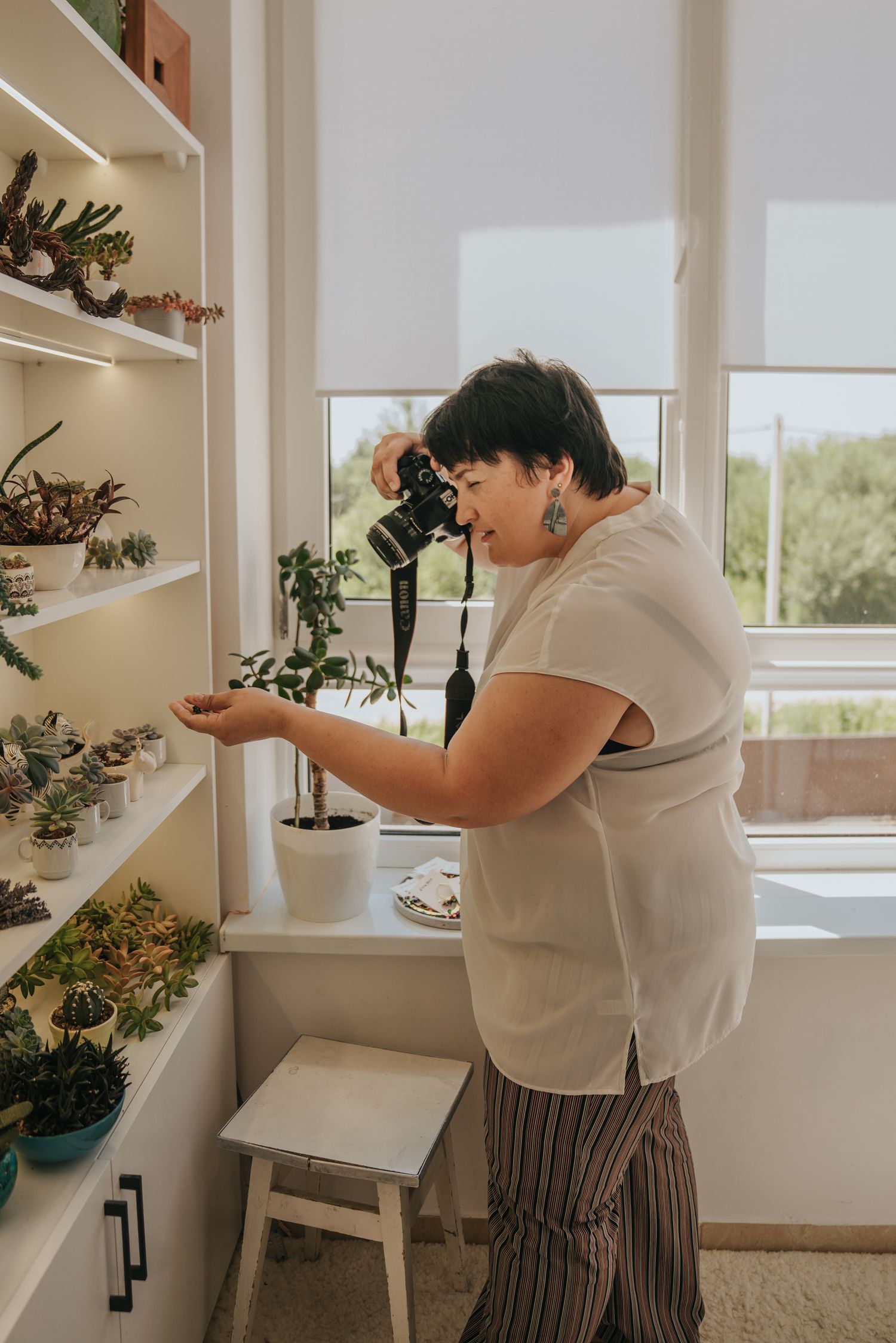 Woman photographer taking close-up photos of succulents on white shelves in a bright home studio.