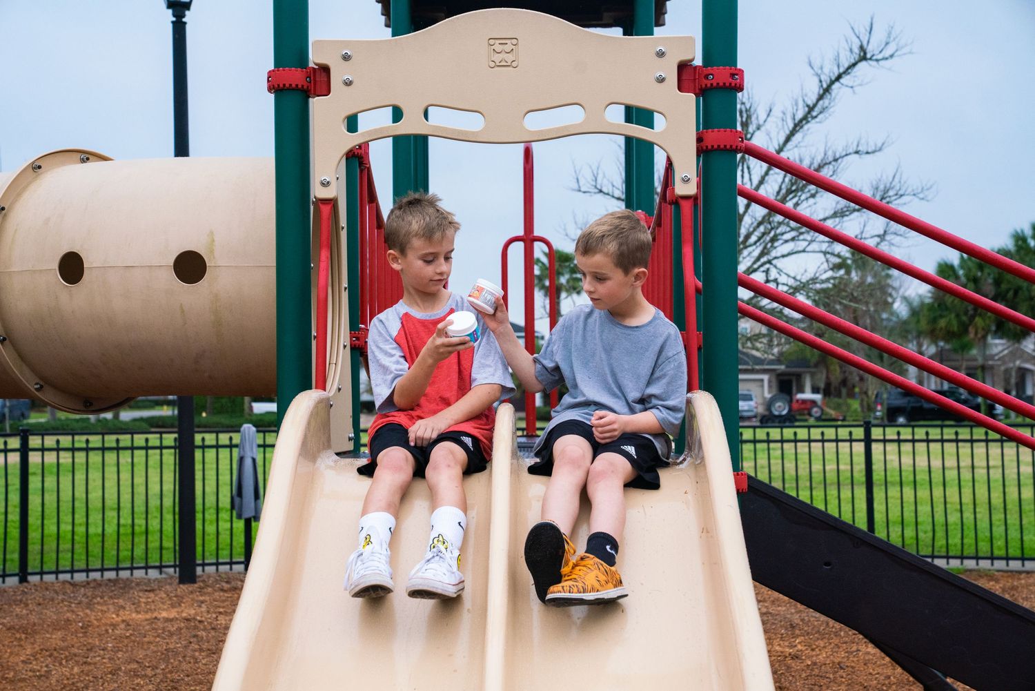 Two boys sitting on playground slides at a park, one holding an ice cream cone on a cloudy day.