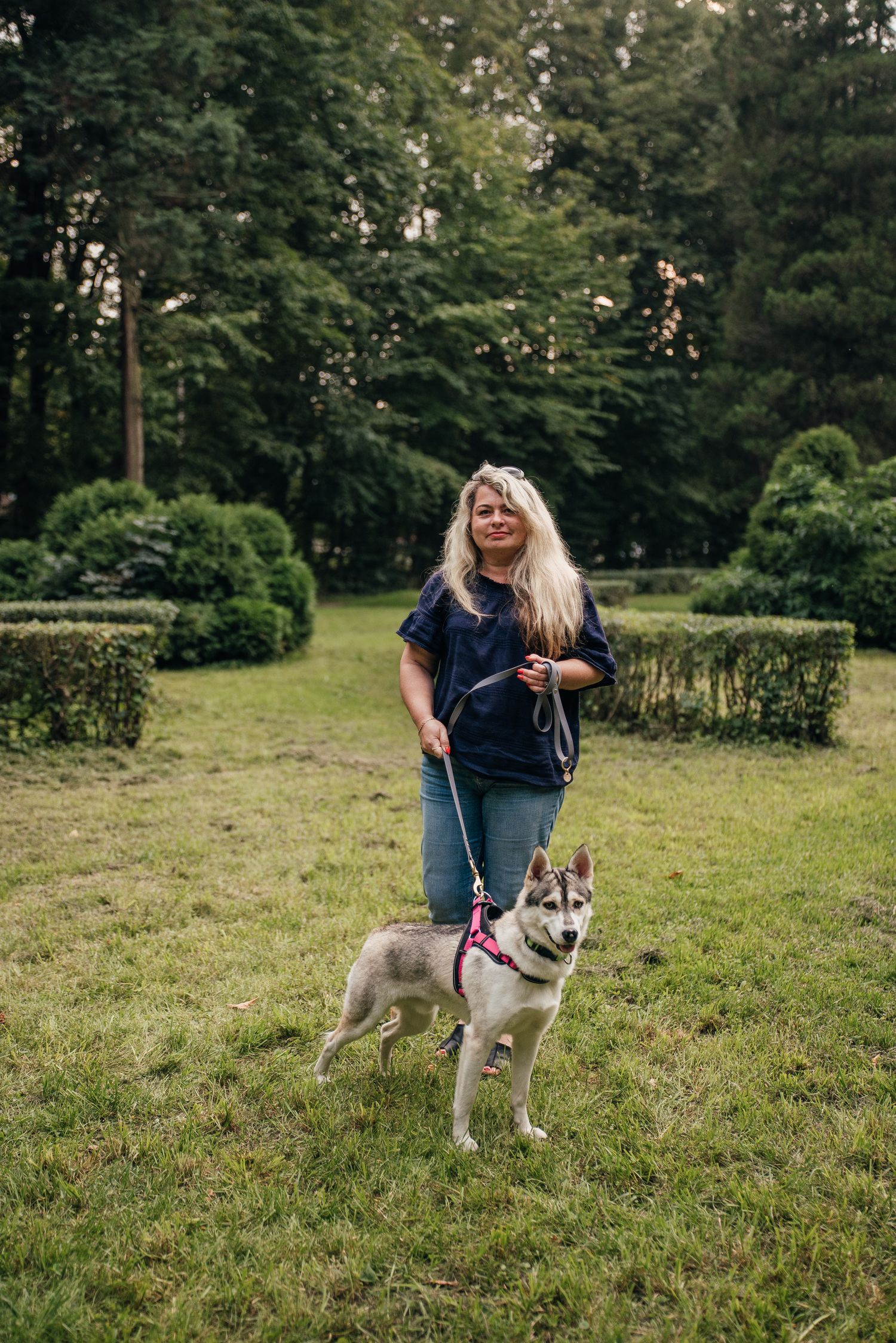 Smiling blonde woman walking a Siberian Husky on a pink leash in a lush green park.