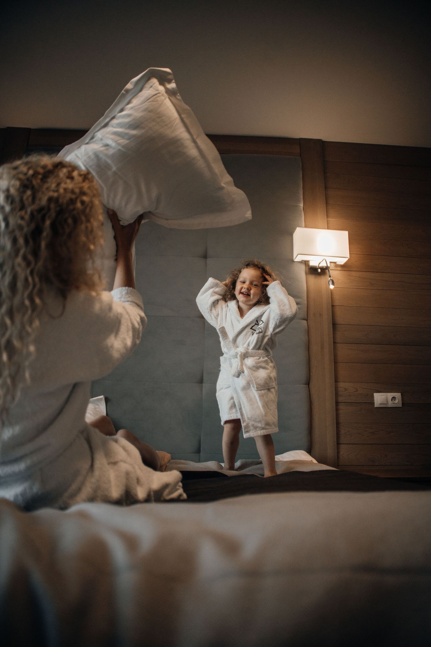 A mother and child in white bathrobes having a fun pillow fight on a hotel bed.
