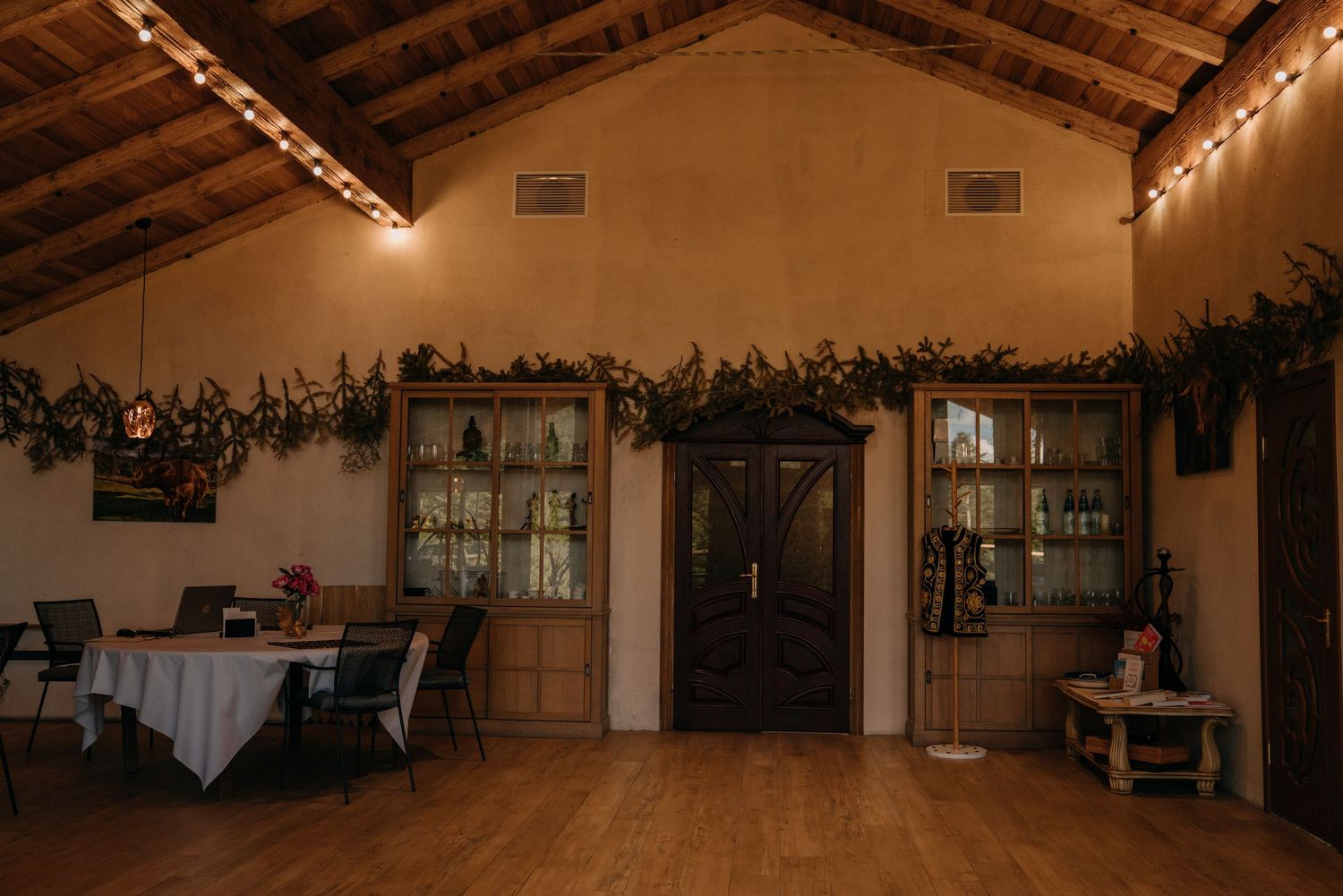 Rustic wedding venue interior with string lights, green garland, wooden doors, and hardwood floors.