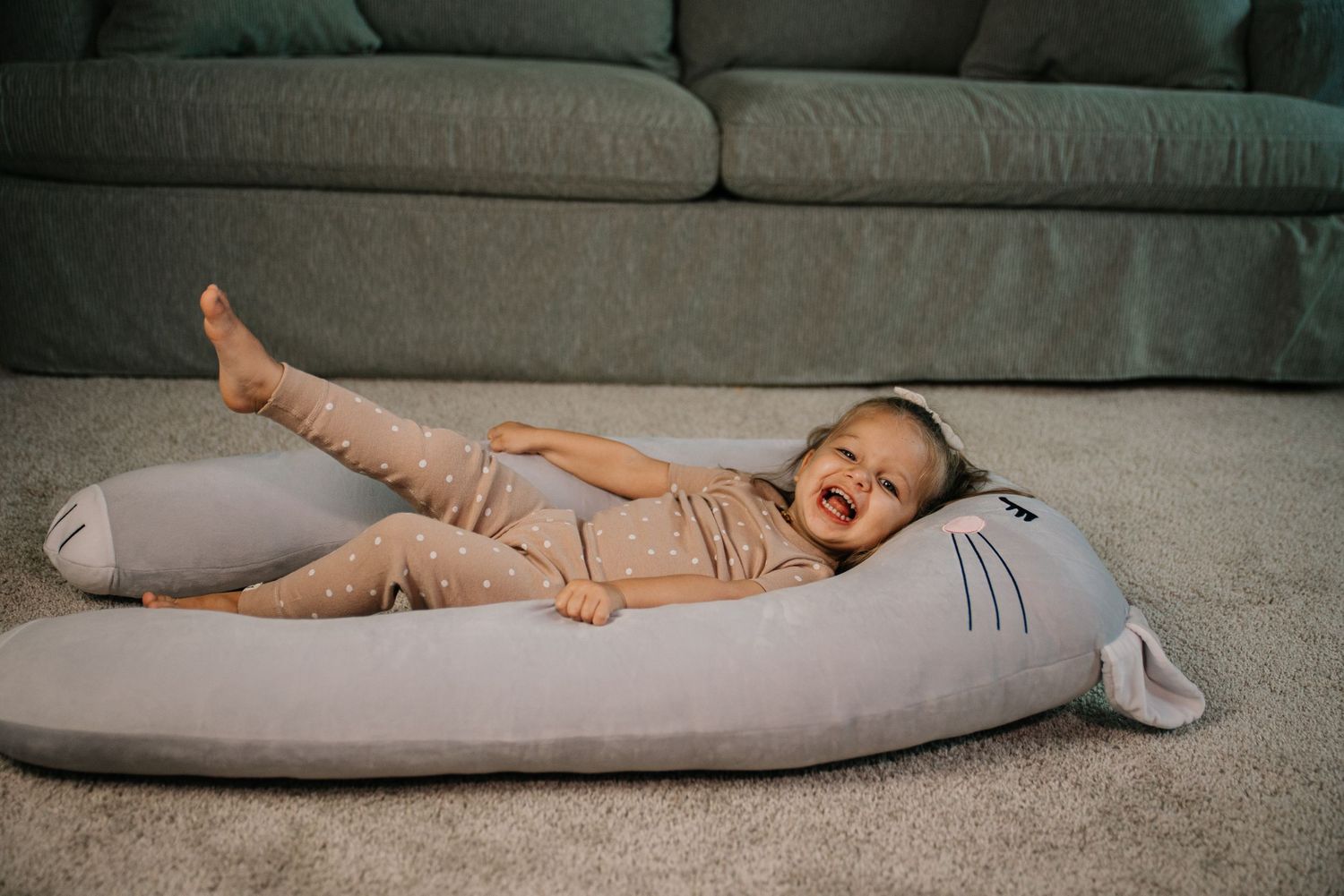 Toddler laughing while lying on a gray shark body pillow near sofa.