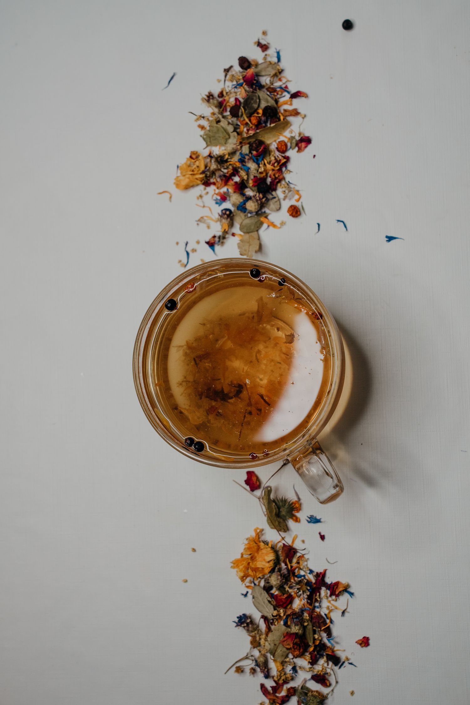 Top view of a glass cup of herbal tea surrounded by colorful dried flower and herb tea blend on white surface.