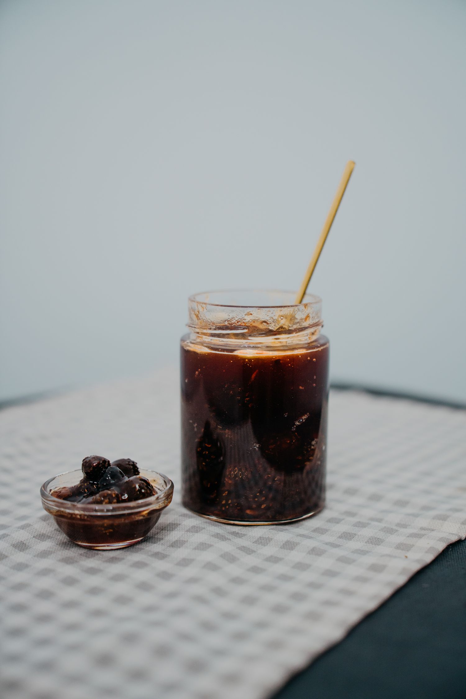 Glass jar of dark blackberry jam with a wooden straw beside a small bowl of berries on checkered cloth.