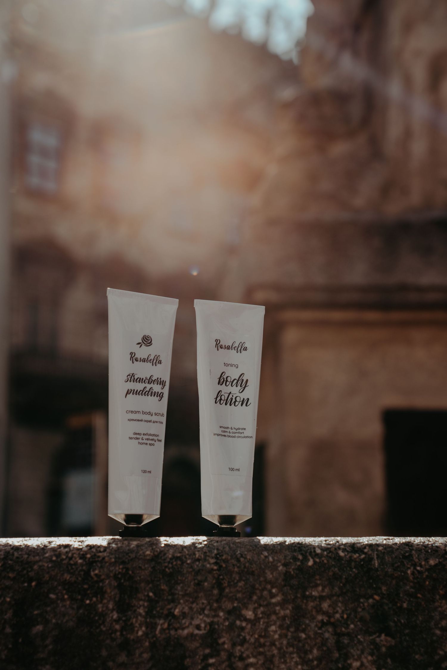 Two Rusaloffa skincare tubes, Strawberry Pudding and Body Lotion, displayed outdoors in warm sunlight.