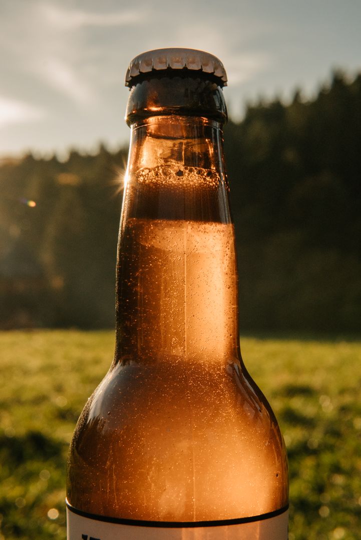 Close-up of a cold beer bottle with condensation, backlit by golden sunset light with green trees in background.