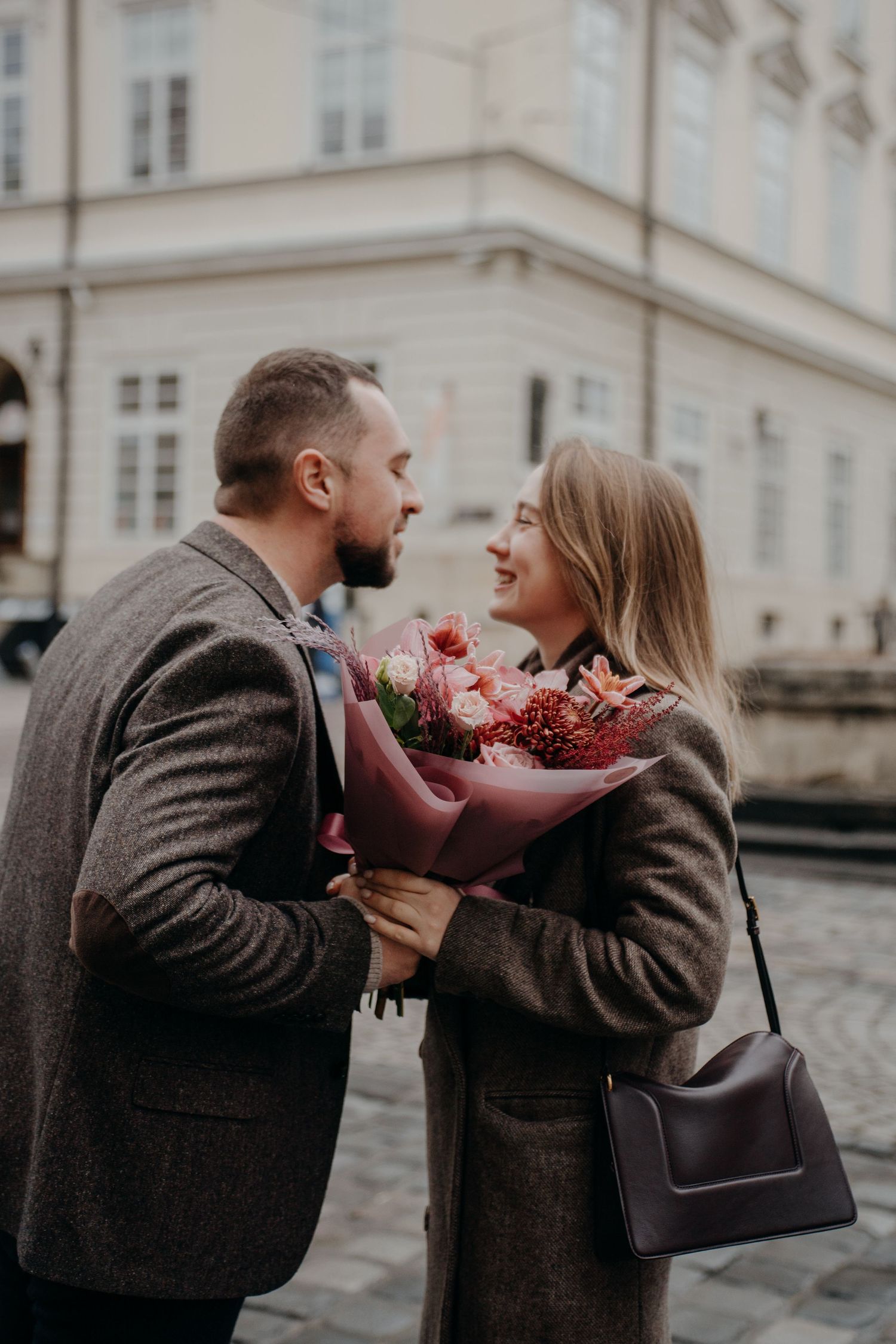 Romantic couple sharing a bouquet of pink flowers on a cobblestone street, smiling at each other in autumn coats.