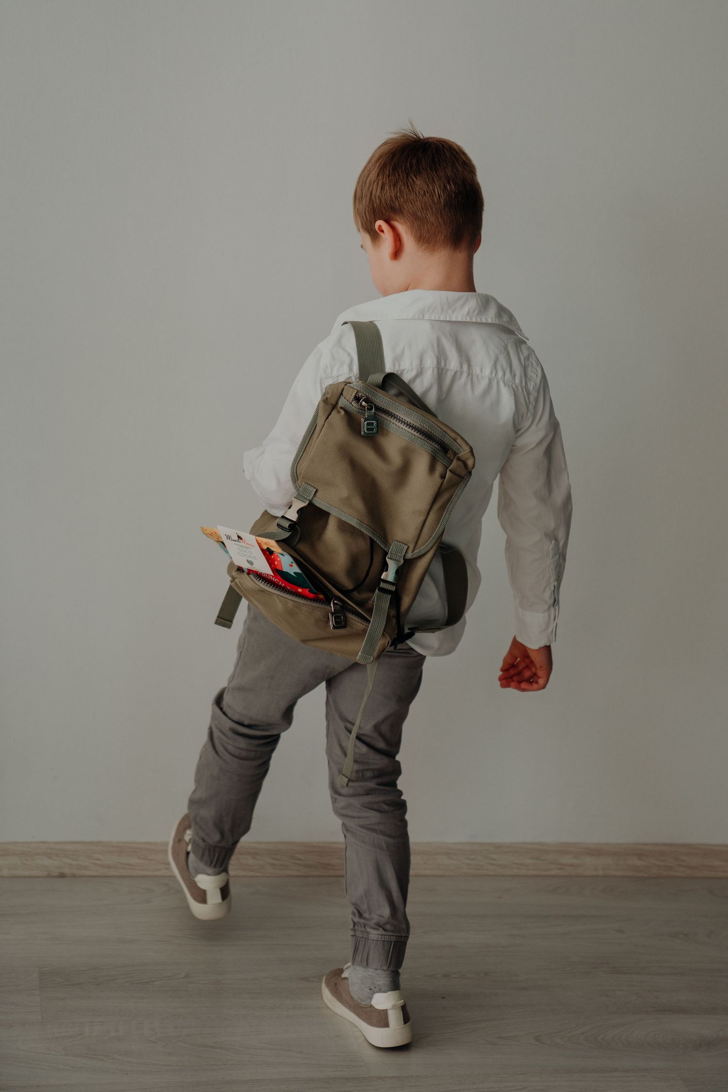 Young boy in white shirt and gray pants walking away with a khaki backpack full of school supplies.