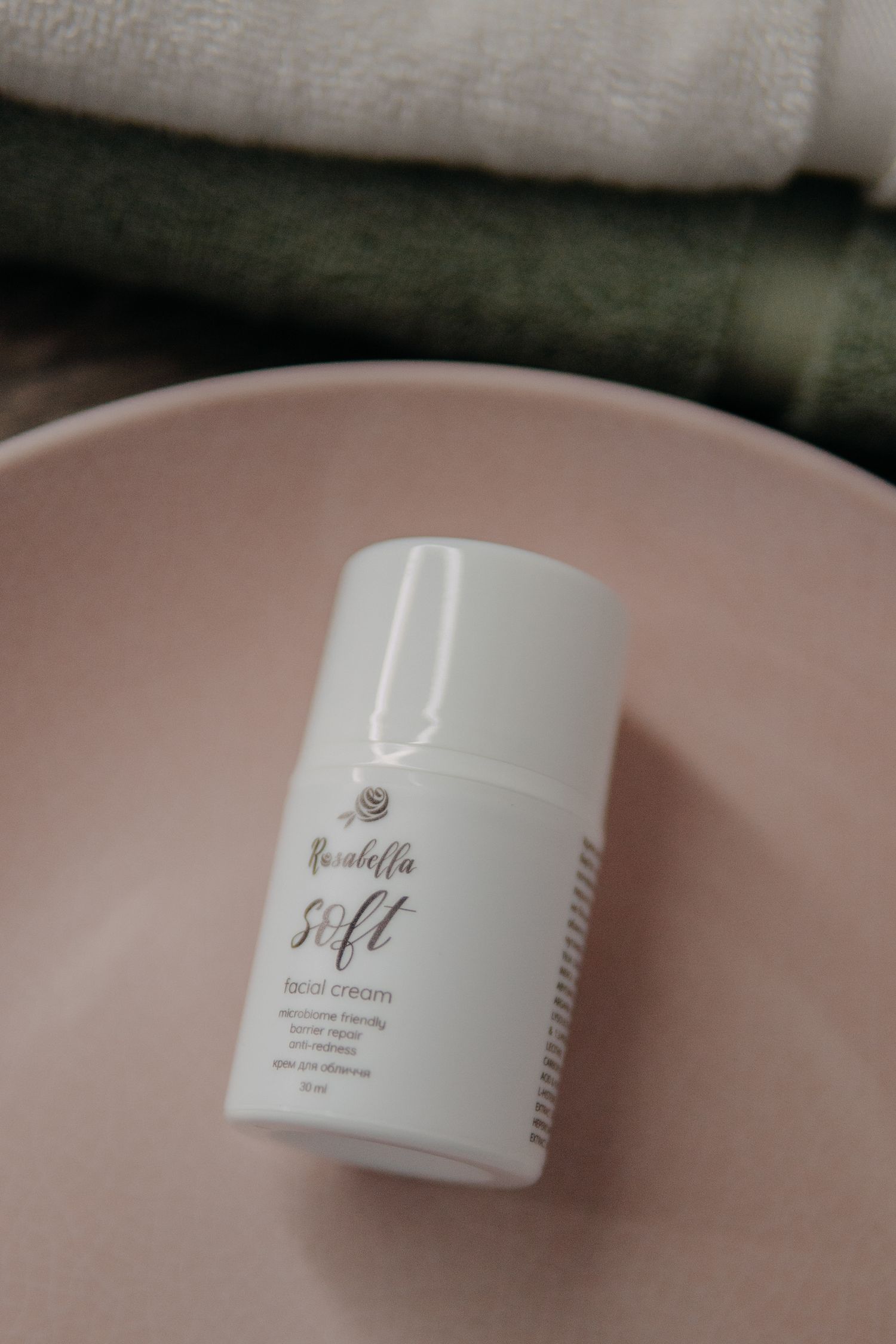 Rosabella Soft facial cream in white pump bottle resting on pink ceramic dish with folded towels in background.
